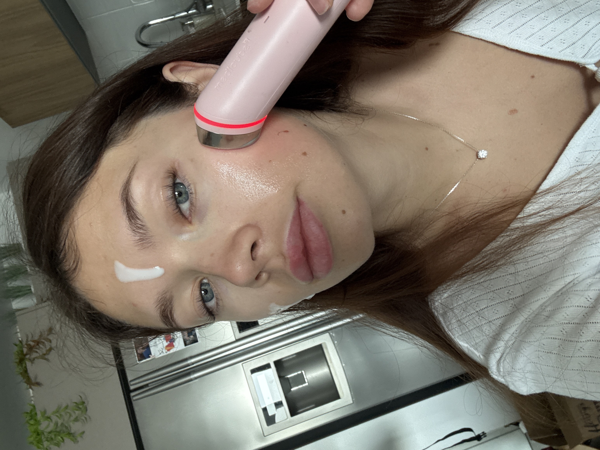 A woman with fair skin, light blue eyes, and dark brown hair is using a pink electronic facial device on her right cheek. A small amount of white cream is visible on her forehead and below her mouth. She is wearing a white, possibly crocheted or knitted, shirt and a delicate silver necklace with a single diamond pendant. The background shows a kitchen interior with stainless steel appliances, including a refrigerator and a faucet. The scene likely depicts a beauty routine in a home setting. There's no specific evidence that points to the city of Givatayim, Israel. The location is inferred from the image.