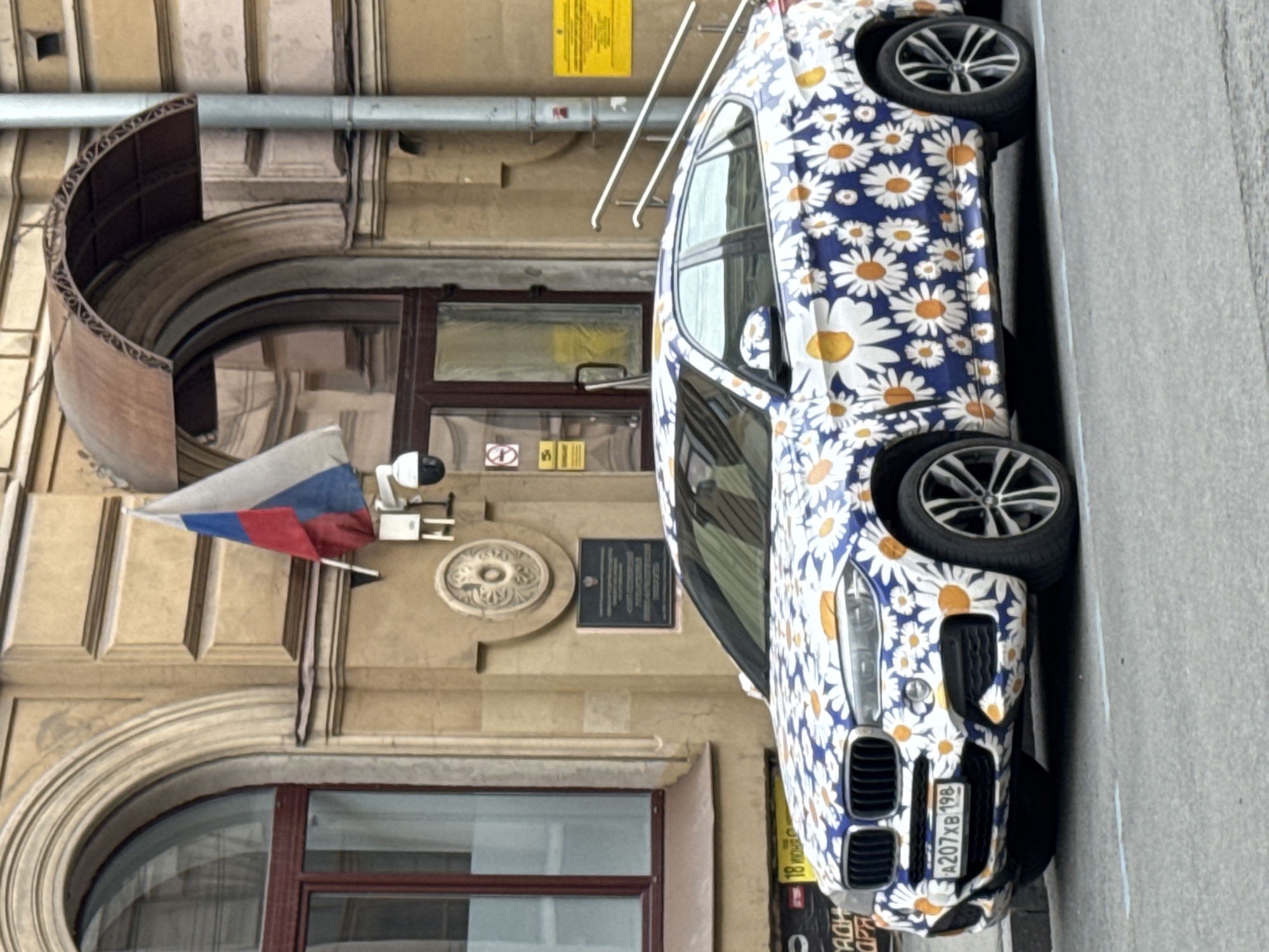 The image shows a building in Saint Petersburg, Russia, with a Russian flag displayed next to the entrance. A car with a distinctive daisy floral pattern is parked on the street next to the building. The license plate of the car is visible. The building features an arched window, an arched doorway with a dark brown door, and a security camera mounted above the doorway. A metal sign or plaque is also visible on the facade of the building. The scene appears to be captured during daylight hours.