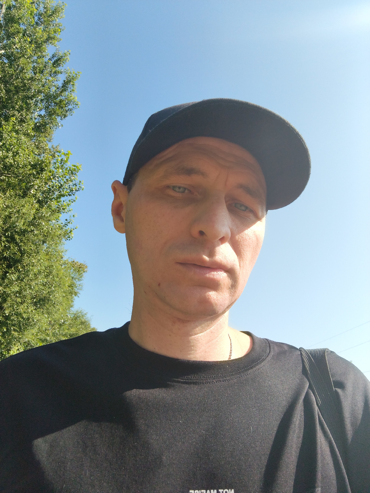 A low-angle, waist-up selfie shows a man wearing a black t-shirt and a black baseball cap, outdoors under a clear blue sky. To the left of the man, a green leafy tree fills the frame. The man's face is partially obscured by the brim of his cap, and he is looking directly at the camera with a neutral expression. The man has fair skin and light-colored eyes. He is wearing a thin silver chain around his neck. A black bag strap is visible over his right shoulder. The scene appears to be set in a park or a rural area. The overall lighting suggests it is daytime.