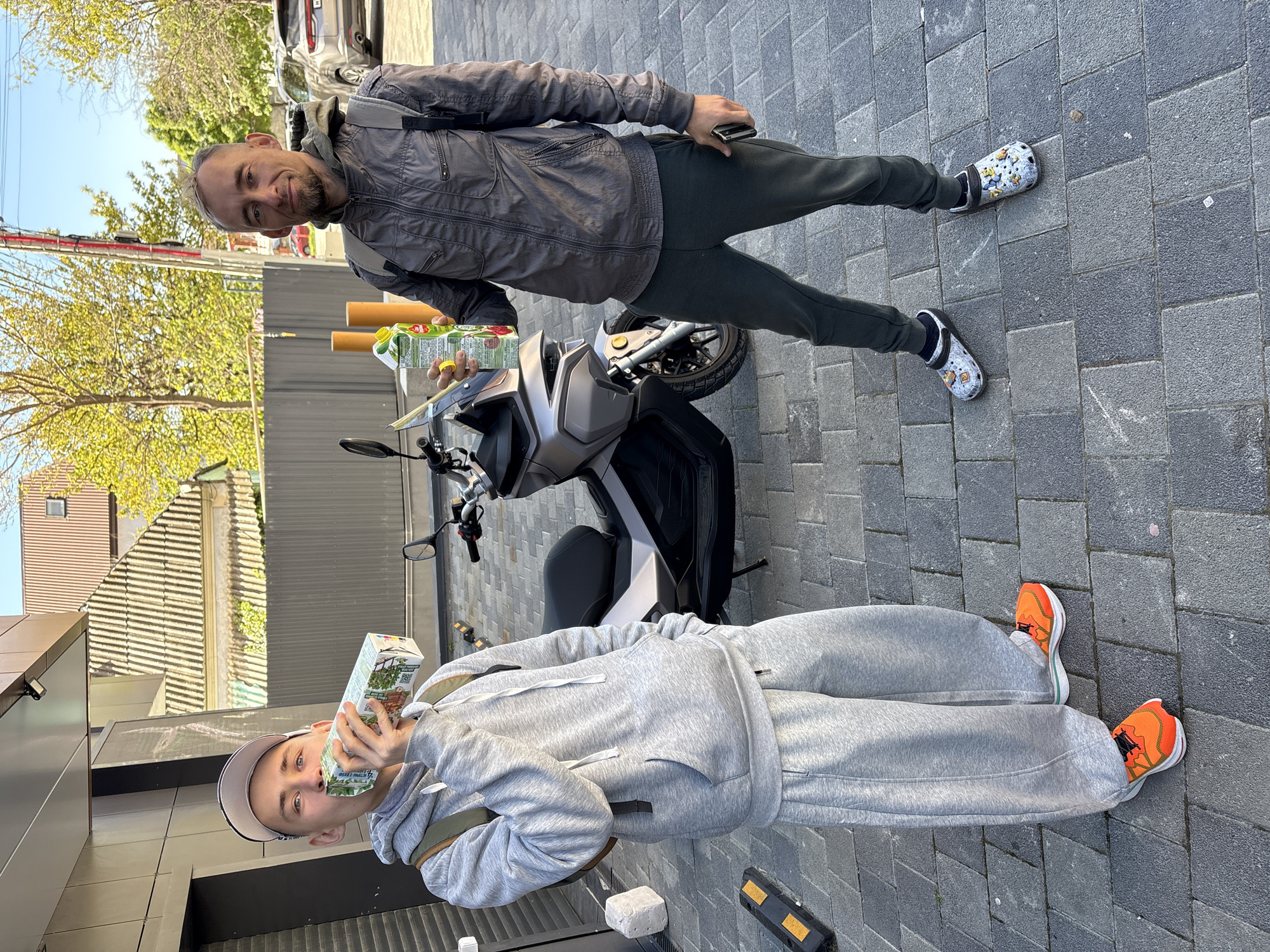 Two men are posing outdoors on a sunny day in Gelendzhik, Russia. The image shows them standing on a grey paved surface, with a scooter parked between them.
On the left side of the frame (viewed vertically), a younger man is wearing a grey tracksuit and a light-colored baseball cap. He is holding a white carton of drink to his face, appearing to take a sip, and looking directly at the camera. He wears bright orange and white sneakers. The drink carton has green text that partially reads "BEZ G...I" and "MEGANO B", along with an illustration of plants or herbs, suggesting a natural beverage.
On the right side, an older man with a short beard is wearing a dark grey jacket, dark grey trousers, and a small backpack. He is smiling warmly at the camera, holding a yellow carton of drink in his left hand and a mobile phone in his right. The carton has partially visible text like "Pripravs K" and an illustration of fruits, possibly apples or pears. He is wearing distinctive white clogs with yellow cartoon-like characters, possibly Minions, on them.
A light-colored scooter or small motorcycle is positioned between the two men. In the background, there are buildings, including one with a brown facade and another with a tiled roof, as well as trees with green foliage under a clear, bright sky, indicating good weather. The ground is made of interlocking grey paver bricks.