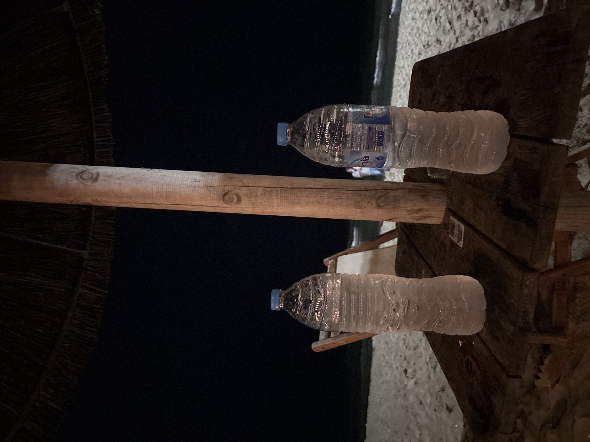The image depicts a nighttime scene on a sandy beach, likely in Varna, Bulgaria. In the foreground, a rustic, dark wooden table is positioned beneath a central wooden pole, which supports a thatched umbrella (partially visible at the top). Two clear plastic water bottles with blue caps, identified as "Ionia" brand and appearing to be 1.5L, stand upright on the table. Both bottles exhibit condensation or frost, indicating they are cold.
In the mid-ground, the sandy beach extends towards the water. A light-colored wooden beach chair is partially visible behind the left water bottle. Faint, indistinct human figures are discernible in the distance, near the waterline. The background features the dark expanse of the Black Sea, with white-capped waves breaking gently on the shore under a completely black night sky. Ambient lighting illuminates the immediate foreground.