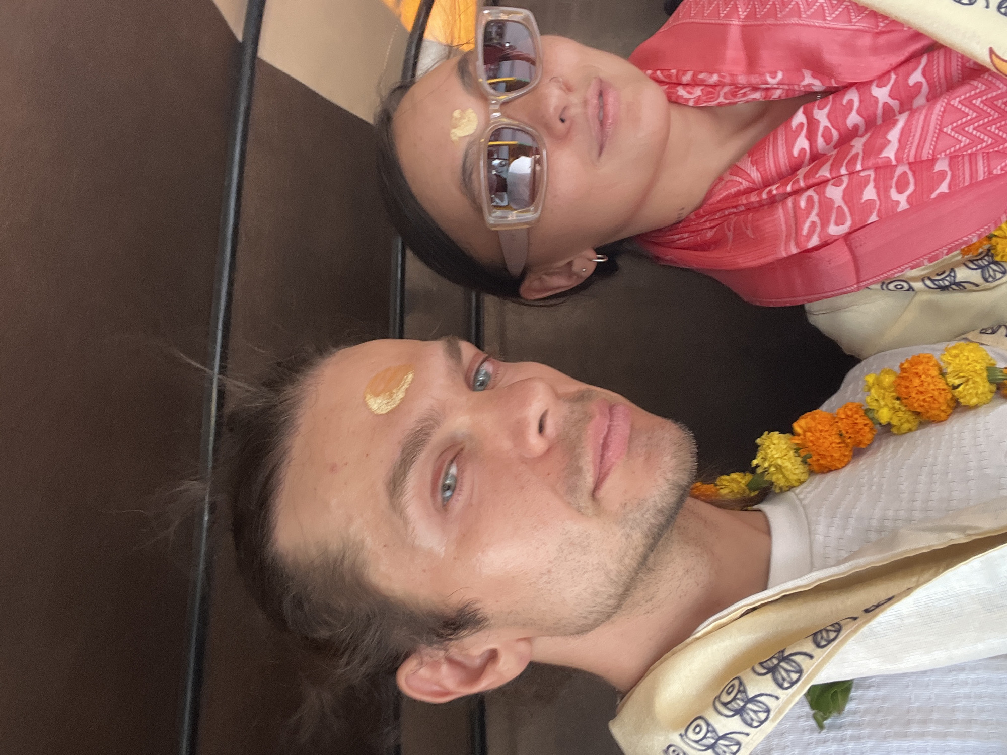 A selfie-style image shows two individuals, a man and a woman, possibly tourists, in Varanasi, India. Both have tilaks on their foreheads and are adorned with flower garlands, likely marigolds. The man has light skin, short brown hair, and blue eyes. He wears a white shirt with a gold-trimmed shawl-like garment. The woman, who is darker-skinned and wears sunglasses, has a pink patterned shawl. They appear to be posing for a photo or video in an indoor setting with dark brown walls.