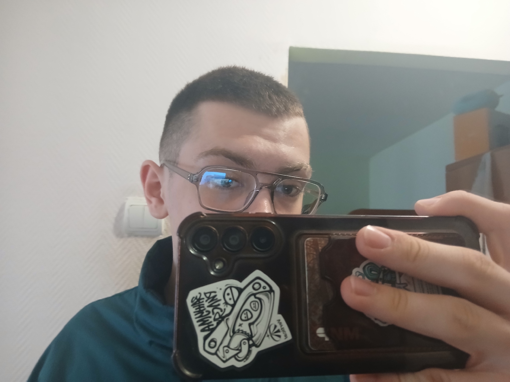 A person with short hair and glasses is holding a black smartphone in front of them, taking a selfie. The phone has stickers on its back. The person is wearing a green shirt with a collar. The background consists of a white wall with an electrical outlet and a doorway leading to another room. Inside that room, some furniture is visible, and the walls are light green.
There is nothing visible in the image that allows the determination of the location in the city of Chaikovsky, Russia.