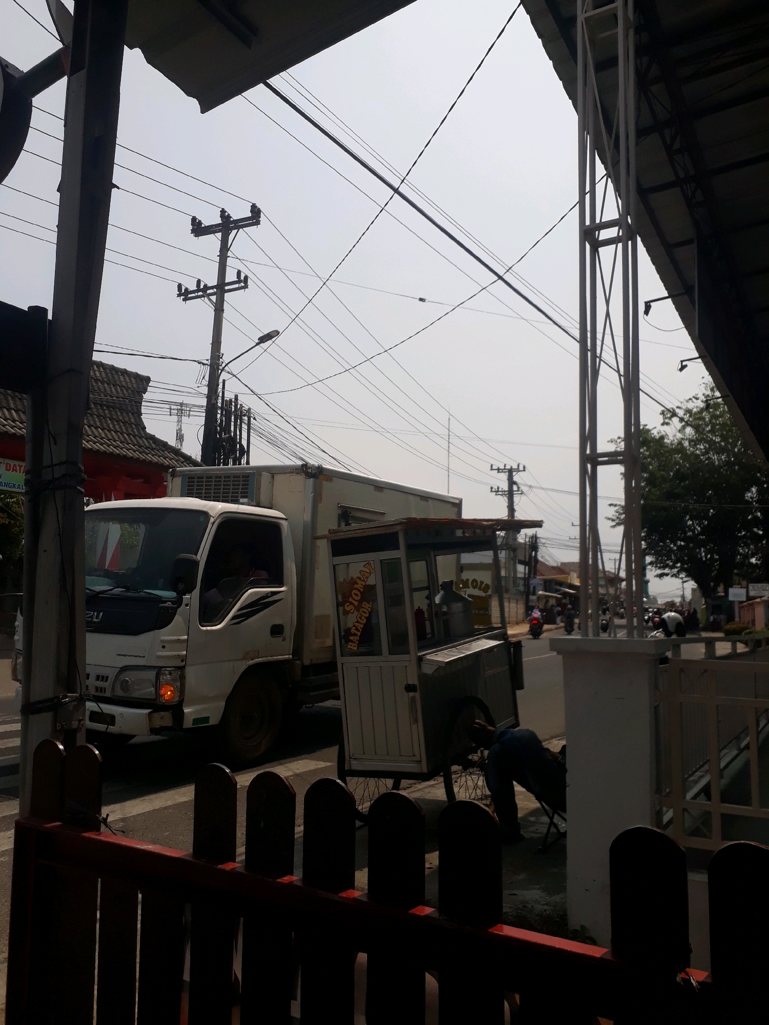 The image captures a street scene in Pangkalpinang, Indonesia, under a hazy sky. In the foreground, a low, dark red fence with vertical wooden slats and a horizontal top rail is visible. Behind the fence, a white truck with a refrigerated cargo box is parked on the left side of the road, its driver's side door slightly ajar. The truck has "ISUZU" branding on the front. To the right of the truck, a food cart with a white and silver exterior is positioned, displaying signage that appears to read "SIOMAY BATAGOR". A person in blue jeans and a dark shirt is leaning against the food cart. In the background, a street with several motorbikes and a few pedestrians can be seen. Utility poles with a complex network of wires crisscross the sky, and various buildings line the street. A white metal structure, possibly part of a covered walkway or building, is prominent on the right side of the image, casting a shadow. The overall impression is of a busy, everyday street scene in an Indonesian city.
