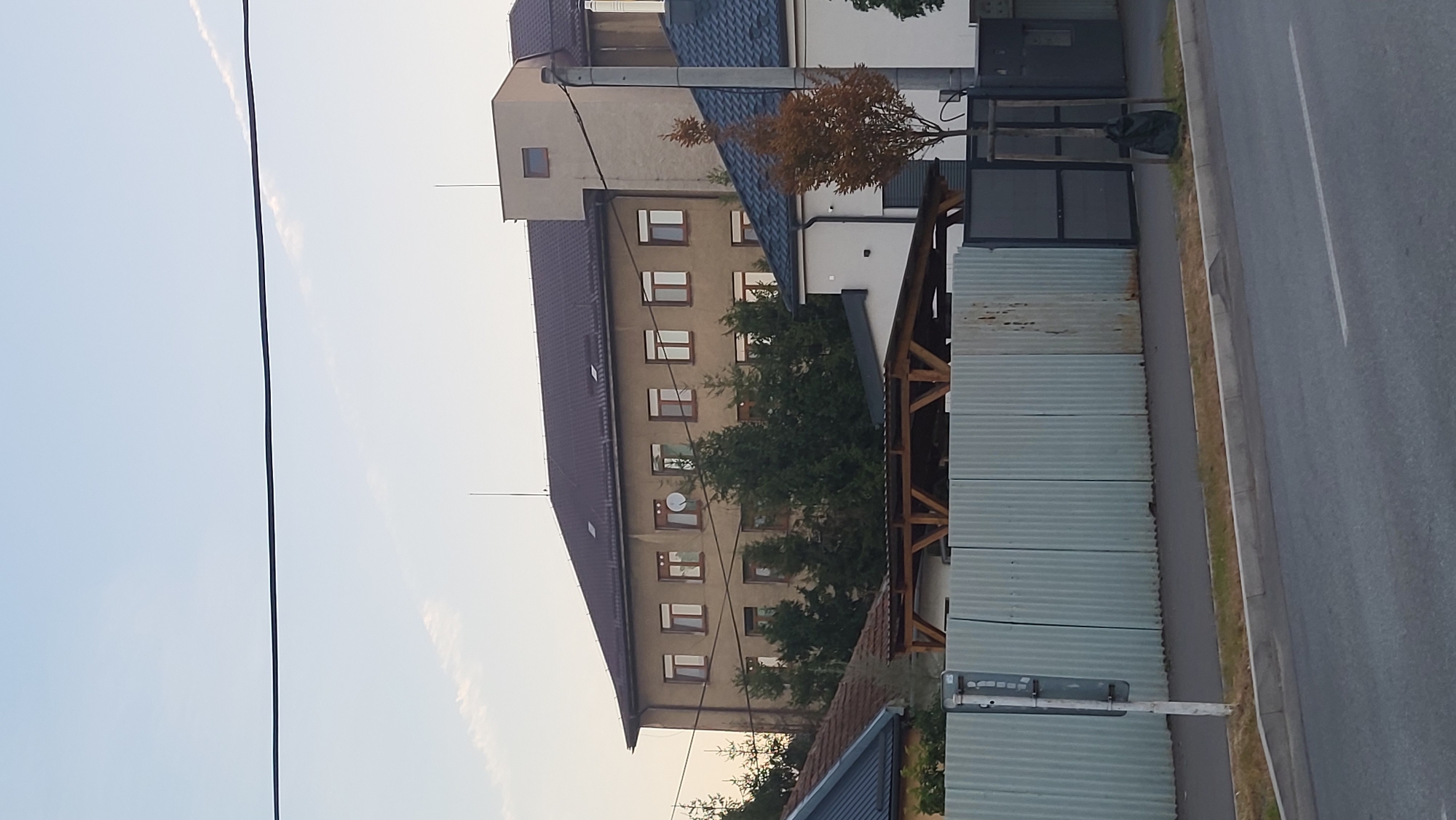 The image depicts a residential building with a brown roof and beige facade, situated in Prešov, Slovakia. The building has multiple windows with wooden frames. In front of the building, there are several evergreen trees and a satellite dish mounted on the facade. A corrugated metal fence runs along the foreground, with a street and sidewalk in front of it. A small tree with brown leaves and a garbage bag are visible near a metal gate. The sky is clear with a few wispy clouds.