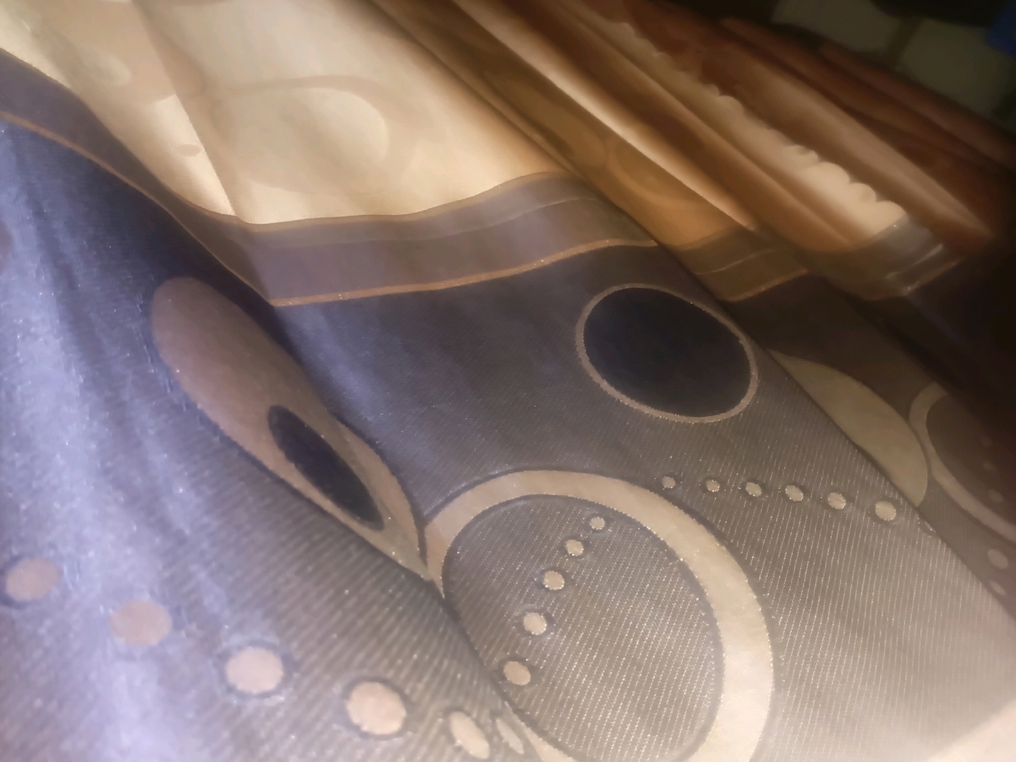 The image shows a close-up of fabric, likely curtains, with a patterned design. The fabric is draped, creating folds and curves. The pattern features concentric circles and rows of small dots in a muted color palette of grays, tans, and browns. The texture of the fabric appears to be slightly shimmering or textured. The lighting is dim, creating shadows and highlighting the contours of the fabric. There is no discernible information about the location in Ol Kalou, Kenya, as the image solely focuses on the textile. No people, actions, or interactions are visible.