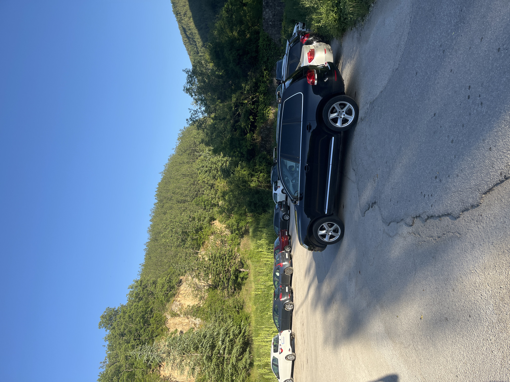 The image depicts a row of parked cars on a road alongside a forested hillside, under a clear blue sky. The cars are various colors and models. The road appears to be asphalt and shows some wear and tear. The hillside is densely covered with green trees and vegetation, and there is a visible rock face in the mid-ground. The image is taken in the vicinity of Blagoevgrad, Bulgaria.