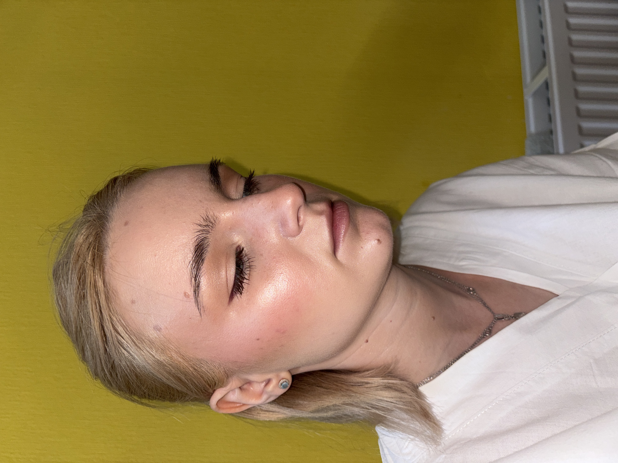A blonde woman with her eyes closed is shown in a close-up, profile view against a bright yellow wall. She has on makeup, including eyeshadow, eyeliner, and blush, and is wearing a white collared shirt and a silver necklace. The woman appears to be indoors, possibly in a room with a radiator visible in the lower right corner. No specific location in Saint Petersburg, Russia, can be determined from the image.