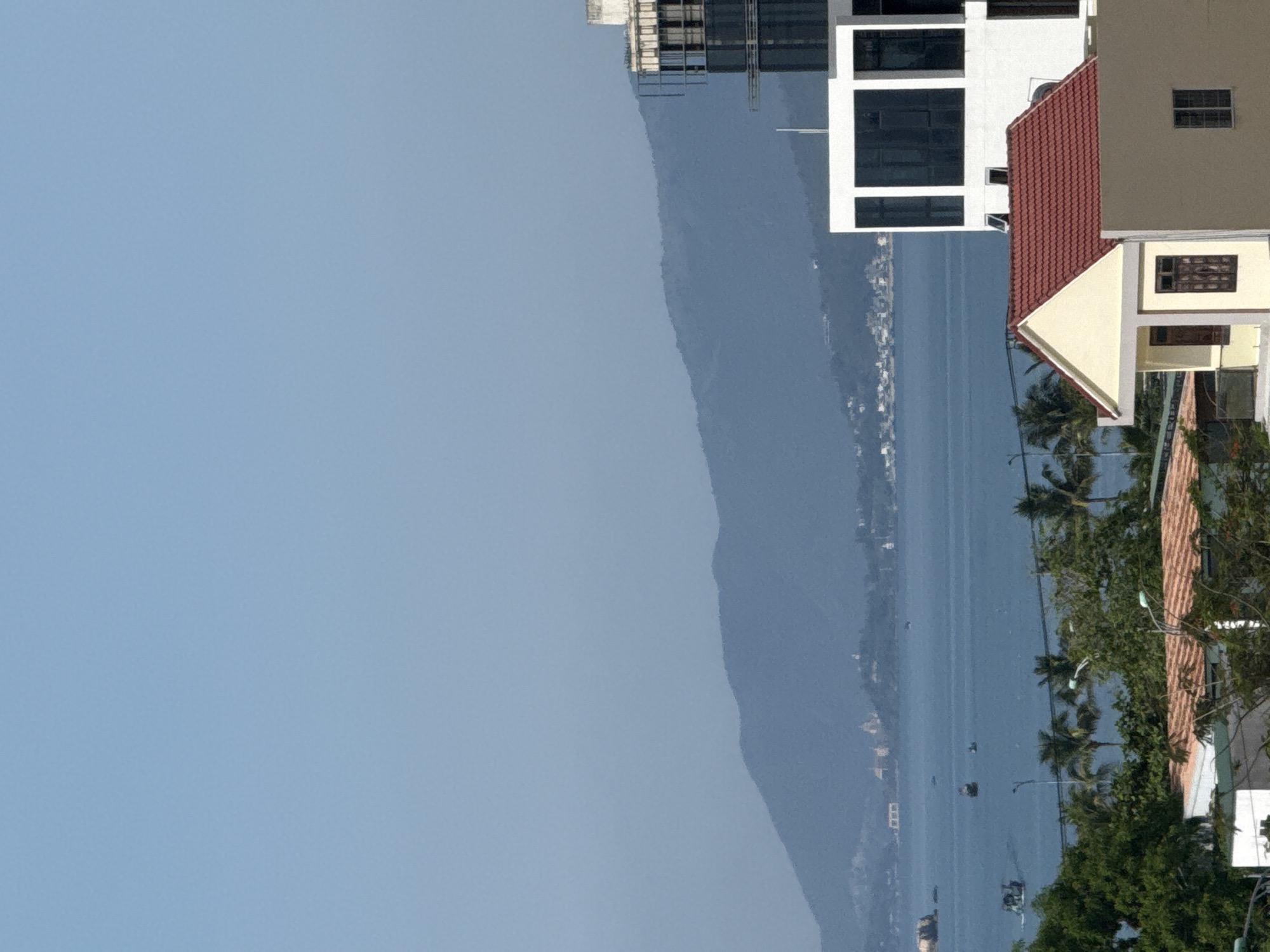 The image captures a panoramic view of the bay in Bac Nha Trang Ward, Vietnam, under a clear blue sky. In the foreground, the roofs of several buildings are visible, including a light yellow house with a red tiled roof and a taller, modern white building with dark windows. Tropical trees, including palm trees, dot the landscape.
The middle ground is dominated by the tranquil, blue water of the bay, dotted with several small boats. In the distance, a range of lush, green mountains forms the backdrop. The shoreline is lined with buildings and vegetation, giving a sense of a coastal city. The overall scene is serene and picturesque, showcasing the natural beauty and urban development of the location.