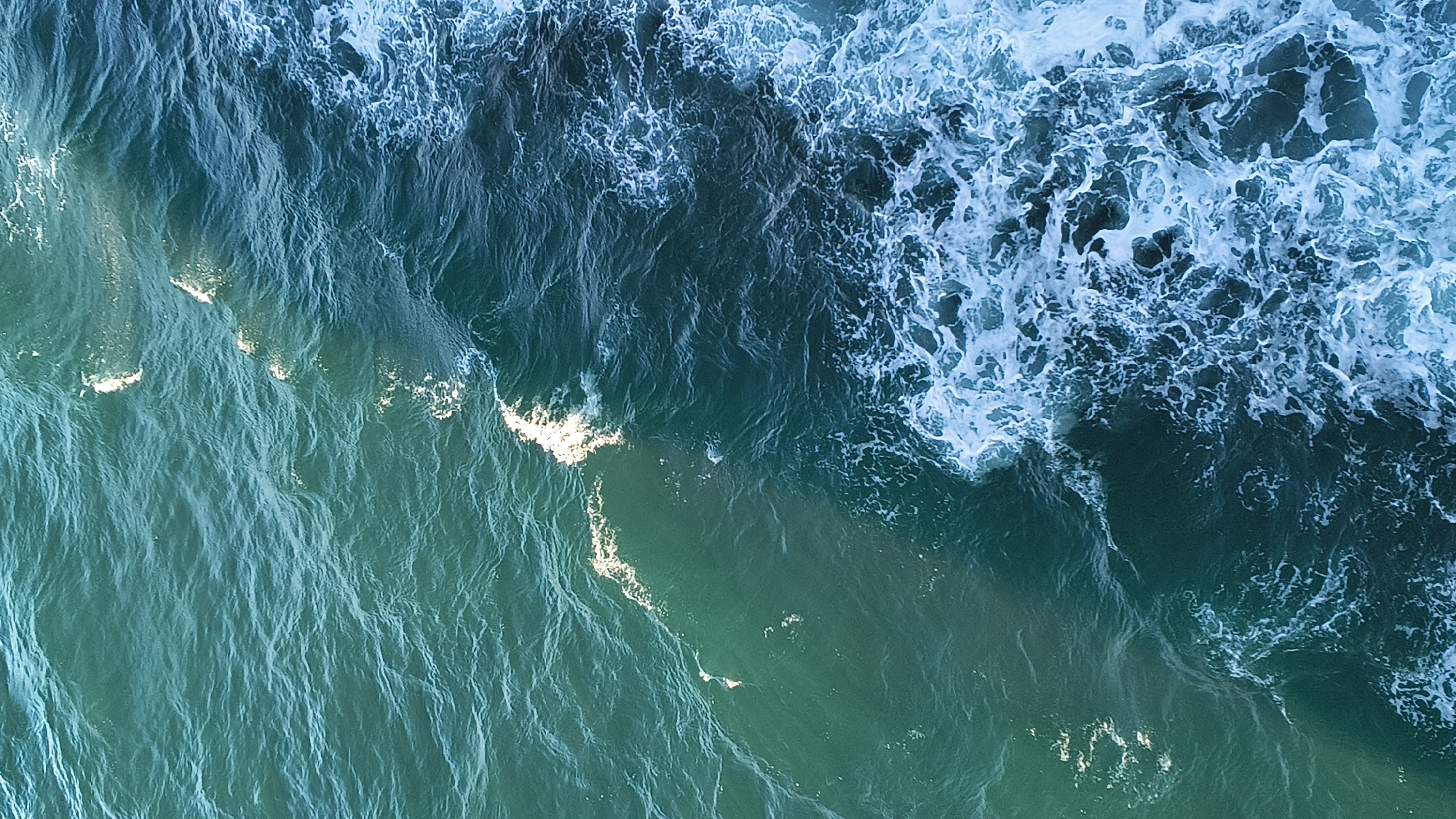 This is an aerial, high-angle, full shot of the ocean. The water is a deep teal color, with lighter turquoise hues where the sunlight hits it. White foam and waves are visible, especially on the right side of the frame, indicating movement and a slightly choppy sea. The texture of the water varies from smooth ripples to churning foam. The scene is devoid of any man-made objects or living beings, presenting a purely natural environment. The location is specified as Unknown, United Kingdom.