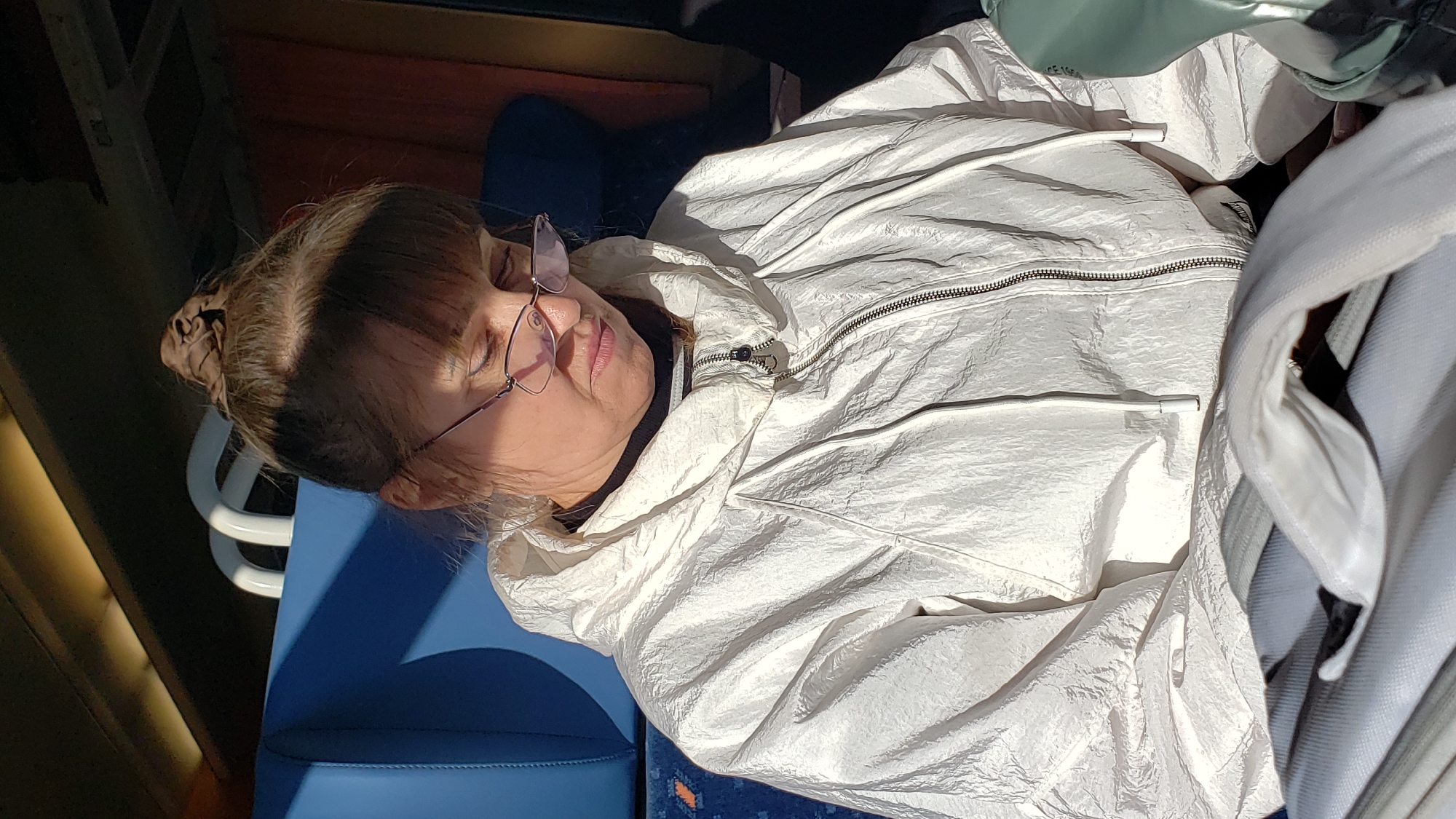 A woman with her hair in a bun is wearing glasses and a white hooded jacket. She is seated on a blue upholstered seat, possibly on a train or bus. The scene appears to be illuminated by natural light, casting shadows on her face and clothing. In the background, there are white arched structures and a red curtain, suggesting an indoor environment. The overall impression is one of a passenger resting or looking down. The location is identified as Važec, Slovakia.