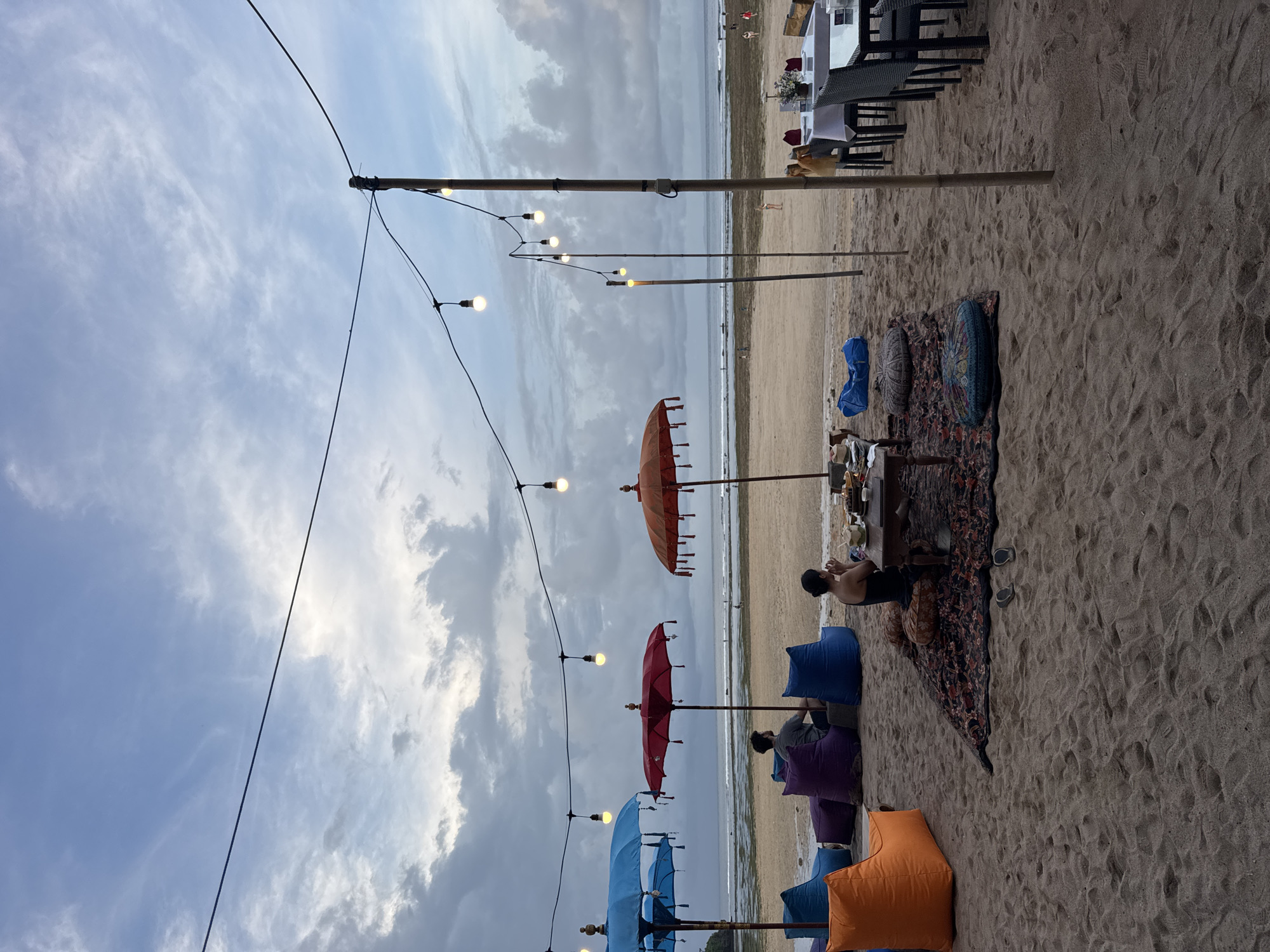 The image shows a beach scene in Kuta Selatan, Indonesia, featuring outdoor seating arrangements. A string of lights extends from the top left to the center of the frame. Below the lights are colorful umbrellas interspersed with cushioned seating areas arranged on the sand. People are seen sitting in these areas, with at least one person appearing to be using a mobile device. A table with items on top is also present. In the background, the ocean meets the shoreline, and further back, there are structures resembling buildings or establishments. Additional tables and chairs are set up closer to these structures. The sky is partially cloudy.