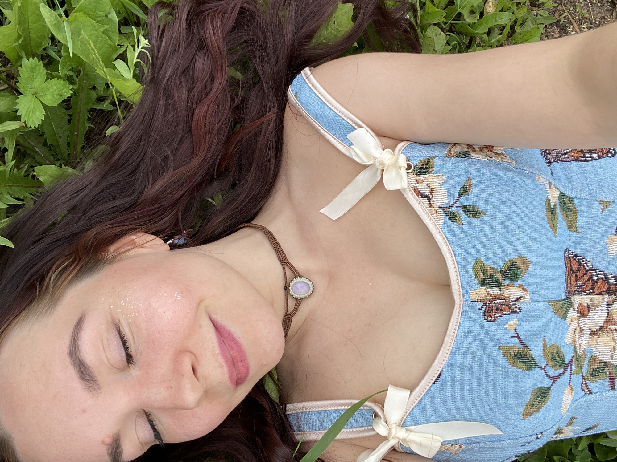 This is a close-up, overhead shot of a young woman lying on her back in the grass. She has long, dark brown hair that is spread out around her head. Her eyes are closed, and she has a slight smile on her face, suggesting a feeling of peace or contentment. There is a subtle dusting of glitter on her cheekbones and forehead, adding a whimsical touch.
She is wearing a blue corset-style top with a floral and butterfly pattern. The top has light pink trim and is adorned with two cream-colored bows on the straps. A macrame necklace with a light purple pendant rests on her collarbone. The background is filled with lush green grass and leaves, indicating an outdoor setting during the daytime. The lighting suggests it's a bright day, possibly late morning or afternoon. The scene evokes a sense of nature and tranquility. No text is visible in the image.