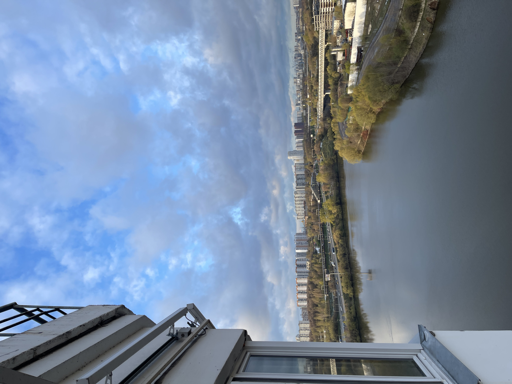 The image captures a panoramic, high-angle view of an urban landscape, likely taken from a tall building overlooking a wide river. The scene is dominated by a partly cloudy sky and the expansive waterway, flanked by developed city banks.
In the upper portion of the frame, the sky displays a mix of bright blue patches and large, soft white and grey clouds, suggesting a moderately overcast day.
Below the sky, a broad river flows, its calm surface reflecting the muted light. Both banks of the river are lined with a combination of trees, which appear to be in early spring with light green or budding foliage, and numerous multi-story buildings, indicating a dense urban or suburban environment. Roads or highways with visible vehicles run parallel to the river on both sides, suggesting active transportation infrastructure. In the distance, more buildings and urban sprawl extend towards the horizon, punctuated by bridges or overpasses.
The immediate foreground features parts of the building from which the photo was taken, including a white railing, a concrete ledge, and a window frame. These elements frame the expansive view beyond.
The time of day appears to be daytime, with soft, diffuse lighting typical of a cloudy day. The trees suggest an early spring season. The setting is an urban waterway, specifically identified as Khimki, Russia. There is no discernible text or specific human activity visible in the image.