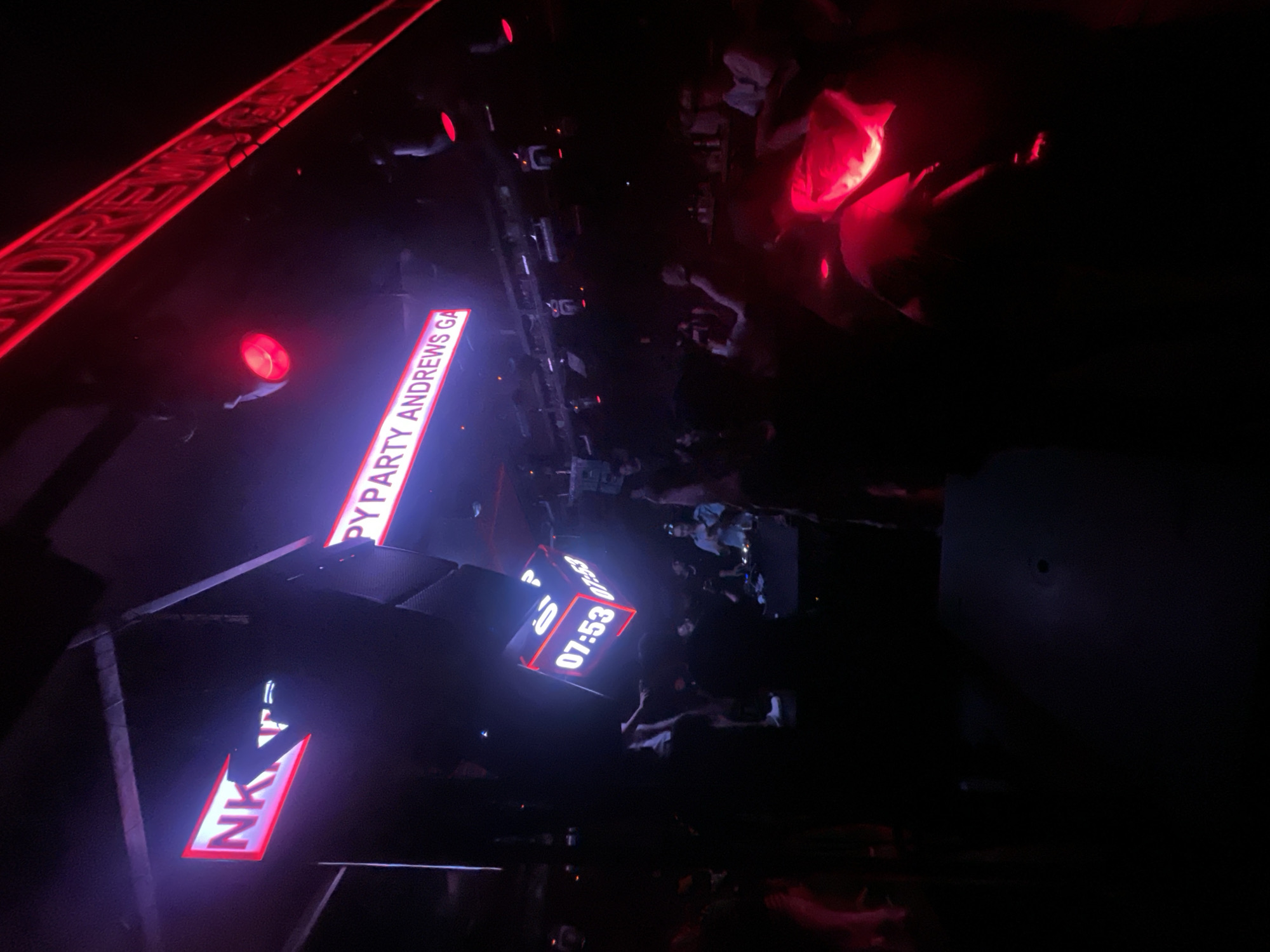 The image captures a dimly lit indoor scene, likely a club or entertainment venue in North Jakarta, Indonesia, illuminated by neon and red lighting. Prominently displayed are three illuminated signs: one with bright pink neon lettering that reads "NK T", another in purple neon text which appears to say "PY PARTY ANDREWS GA", and a third diagonal red neon sign with text partially obscured but recognizable as "DREWS GA". A cube-shaped digital display suspended in the air shows "07:53" on multiple faces. The foreground and mid-ground are filled with silhouettes of people, some appearing to be standing, others seated, and some interacting. Red lighting casts a glow on some of the figures and surfaces. The overall atmosphere suggests a lively social gathering or event.