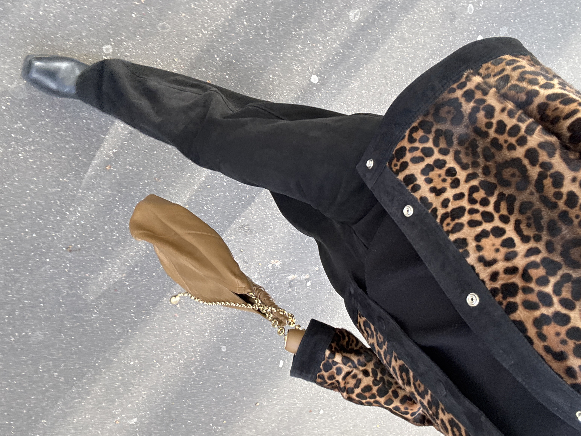 The image displays a high-angle, top-down view of an individual walking on a speckled light grey paved surface in Paris, France. The person is wearing a brown and black leopard print jacket with black trim and silver-colored snap buttons, over a black top and black trousers. A single black, square-toed shoe is visible on the left foot. The individual's right hand holds a light brown, soft-material handbag featuring a gold-colored metal chain strap.