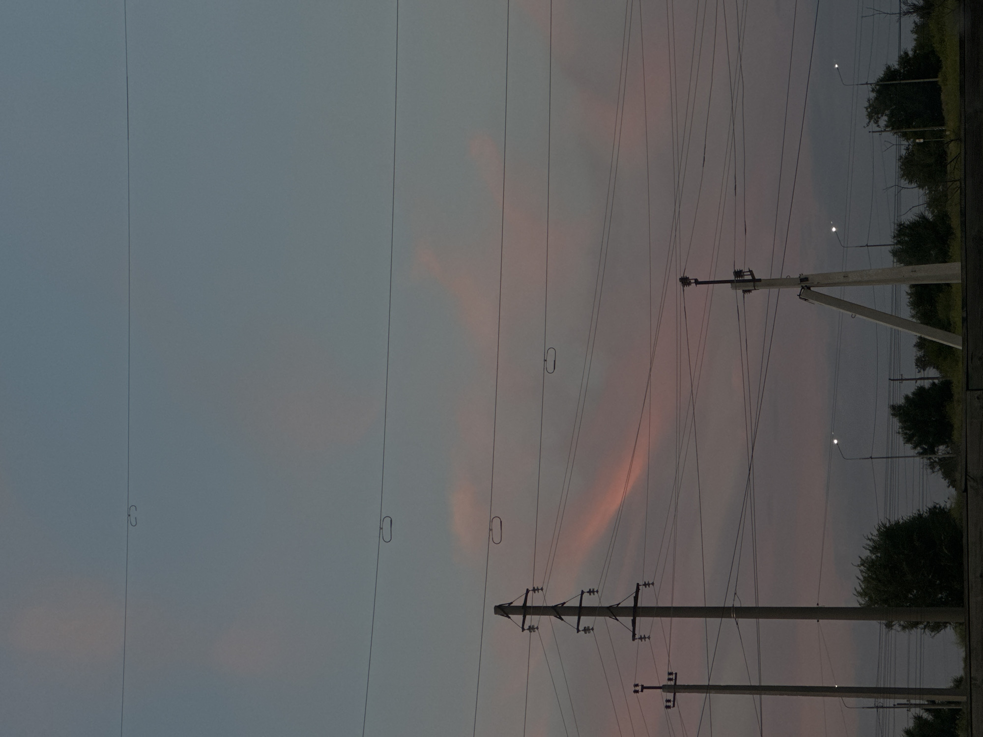 This is a photograph of a twilight sky filled with power lines and silhouetted trees. The sky exhibits hues of gray and pink, suggesting the end of the day. Several power poles and numerous wires crisscross the frame, creating a dense network. In the distance, streetlights are visible, indicating the presence of a road or populated area. Trees line the horizon on the right side of the image, their dark foliage contrasting with the lighter sky. The overall mood is serene and somewhat somber, perhaps reflecting the transition from day to night. There is no visible text in the image.