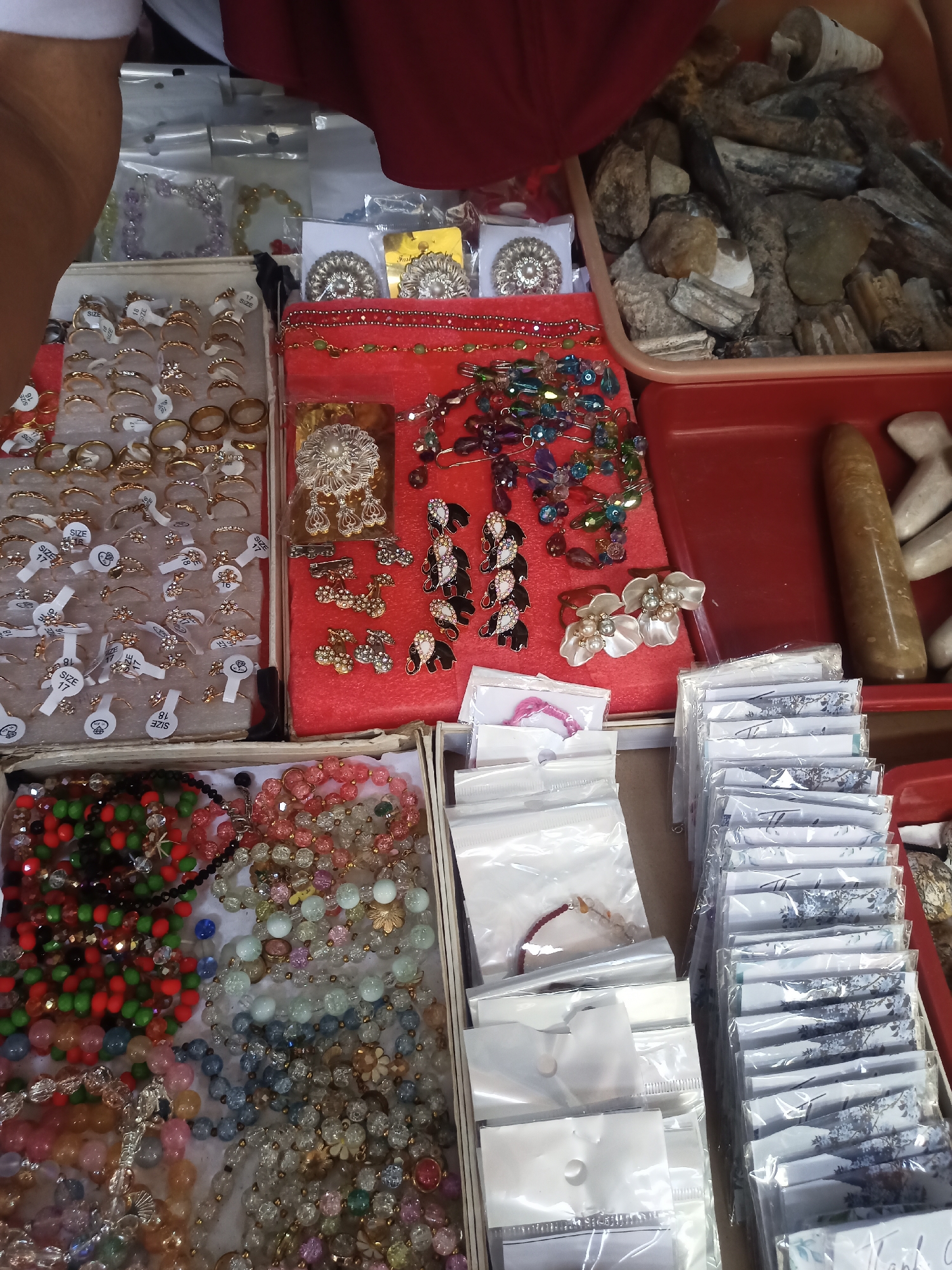 The image displays a street market stall in Gondangrejo, Indonesia, with various jewelry items and natural materials for sale. In the foreground, there are several boxes filled with an assortment of beaded necklaces and bracelets in various colors, including red, green, pink, blue, and clear beads. To the right of the beaded items, a stack of individually packaged greeting cards or small prints with floral designs is visible.
Moving towards the center of the image, a red velvet display holds an assortment of rings, brooches, and earrings. The rings are mostly gold-toned with some featuring clear or colored stones. The brooches and earrings appear to be decorative, with some shaped like elephants and others adorned with pearls and stones. Further to the right, a red plastic tray contains a collection of rough, natural stones and what appear to be fossilized bone fragments. A single, elongated, smooth, brown object, possibly a stone or polished artifact, rests on the edge of this tray.
In the upper left section of the image, more rings are displayed on white cards in a divided box. A person's arm and shoulder, wearing a red garment, are partially visible in the top portion of the frame, indicating their presence behind the stall. The overall scene suggests a busy marketplace environment where a variety of handcrafted and natural items are being offered for sale.