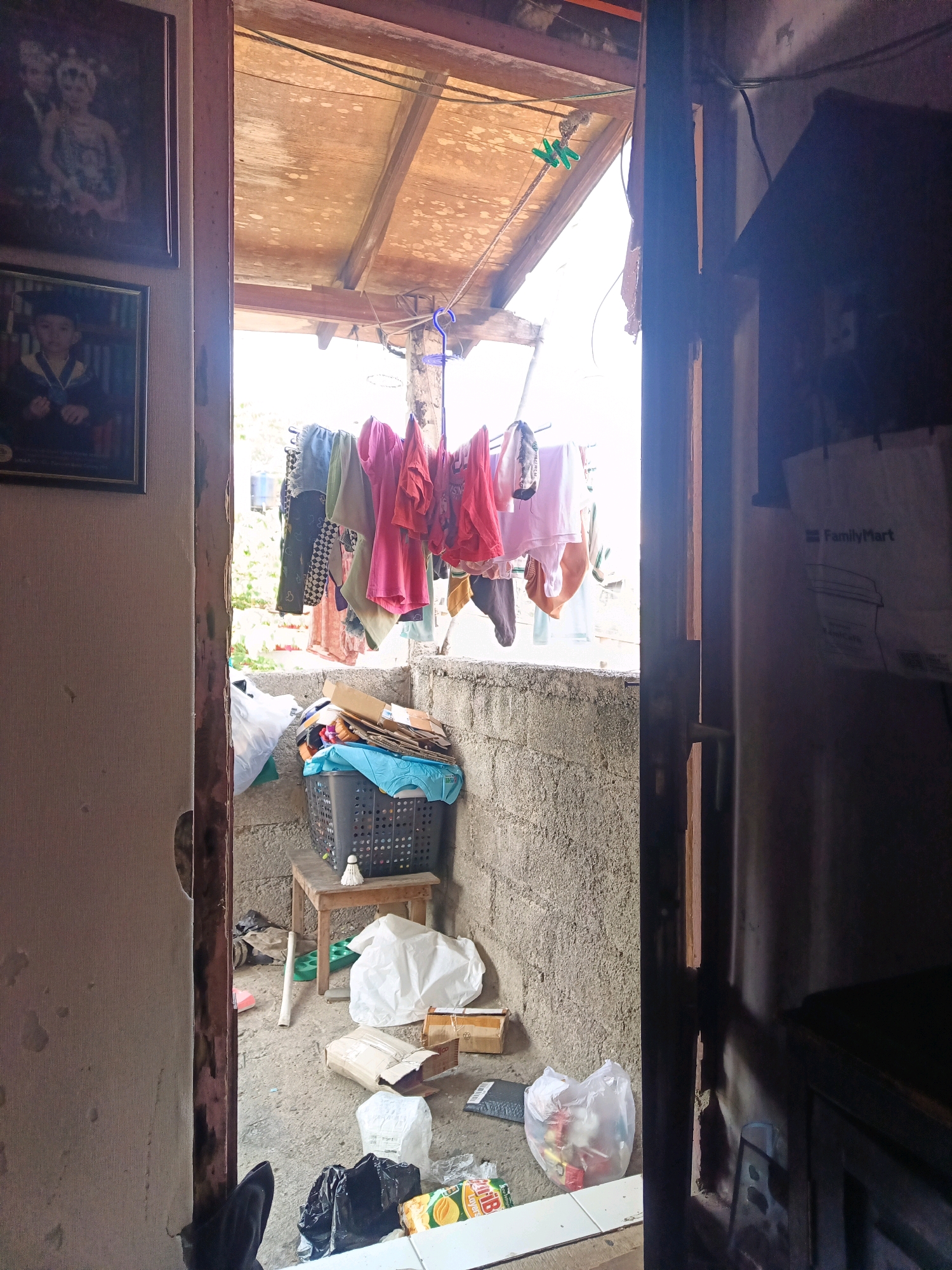 The image depicts a cluttered outdoor area behind a doorway, likely in a residential setting in South Jakarta, Indonesia. The scene is characterized by a variety of items scattered on the ground and suspended from lines.
In the foreground, partially visible on the left, is a wall with two framed pictures hanging on it. The upper frame contains a picture of two people in formal attire, possibly a wedding photo. Below it, another frame shows a child in academic regalia. The doorway itself is open, revealing the space beyond.
The area visible through the doorway appears to be a makeshift laundry area and storage space. Clothes are hanging on a clothesline, some appearing to be brightly colored. A black laundry basket is filled with more clothes and other items, placed on a small wooden stool. Various bags, including white plastic bags and cardboard boxes, are scattered on the concrete ground. A white badminton shuttlecock rests on the stool.
The background shows a rough concrete wall and a sloped, wooden roof structure. Natural light streams in from the right, casting shadows and highlighting the dust and disarray. A black trash bag is prominently placed in the lower foreground on the right, near a plastic bag containing red and yellow items, possibly food. A grocery bag with "FamilyMart" branding is visible on the right side of the image, suggesting proximity to a store or its merchandise. The overall impression is one of a utilitarian and somewhat unkempt outdoor living or workspace.