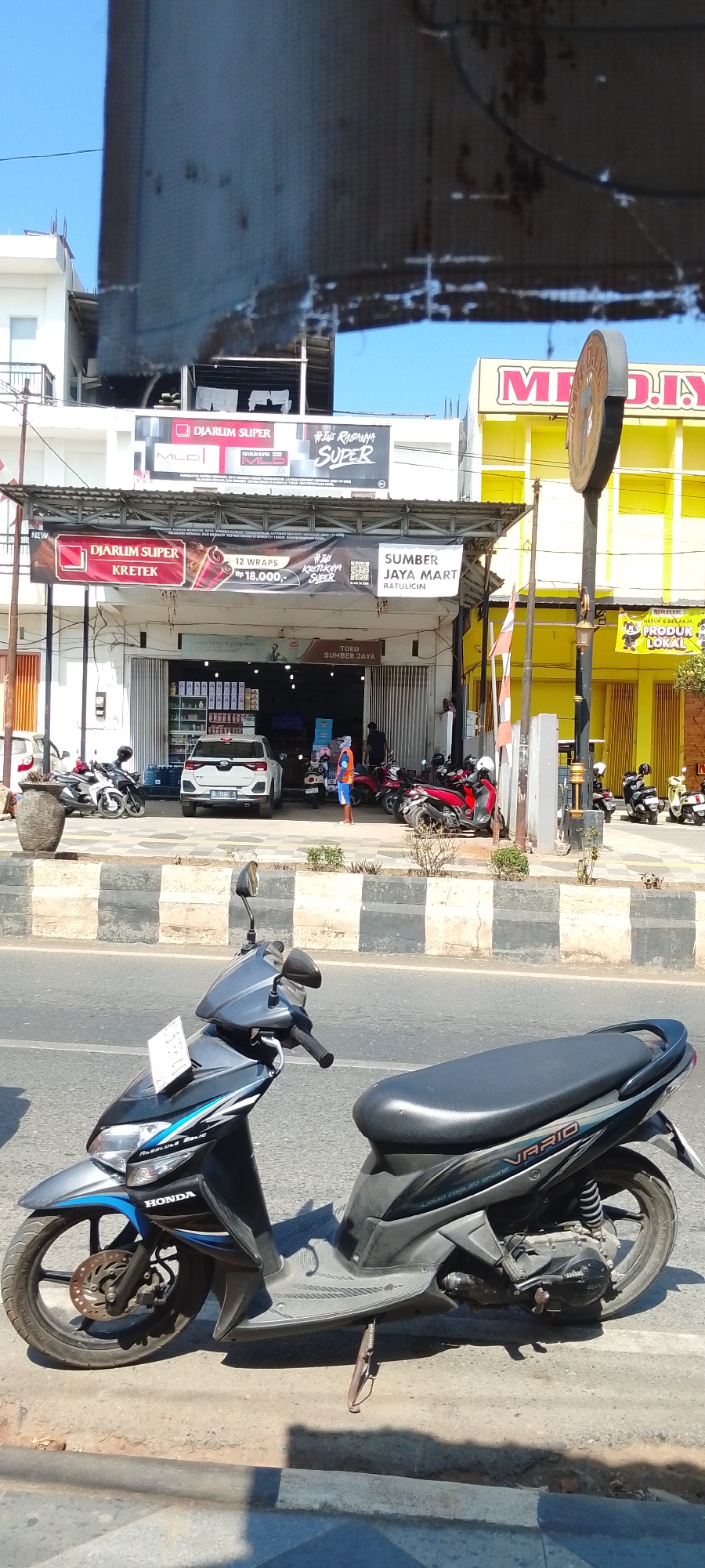 The image depicts a street scene in Batulicin, Indonesia, on a sunny day. In the foreground, a black and blue Honda Vario scooter is parked on the side of the road. Behind the scooter, across the street and sidewalk, is a row of commercial establishments. One building features a yellow facade with prominent red lettering that reads "MITRA DIY". Next to it, a store with a gray awning and an open entrance displays various goods inside. Overhead banners announce "DIARUM SUPER KRETEK" and "SUMBER JAYA MART RATULICIN". Several motorcycles are parked in front of the stores, and a white SUV is visible in the open doorway of one establishment. A person wearing an orange vest and blue shorts stands near the entrance of a store. The street has a black and white striped median barrier. The sky is clear blue, and shadows are cast on the ground, indicating the time of day is likely late morning or early afternoon.
