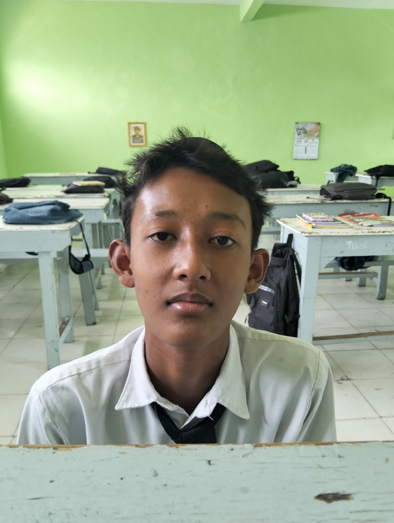 A boy is photographed from the chest up in a classroom setting. He is wearing a white collared shirt with a black tie. The boy has dark, spiky hair, brown eyes, and his skin appears oily. The background is a bright green wall with two framed items hanging on it. In front of the boy are several desks, some with bags and books on them. The floor is tiled. The image is likely taken in Kranji, Indonesia, judging by the provided context.