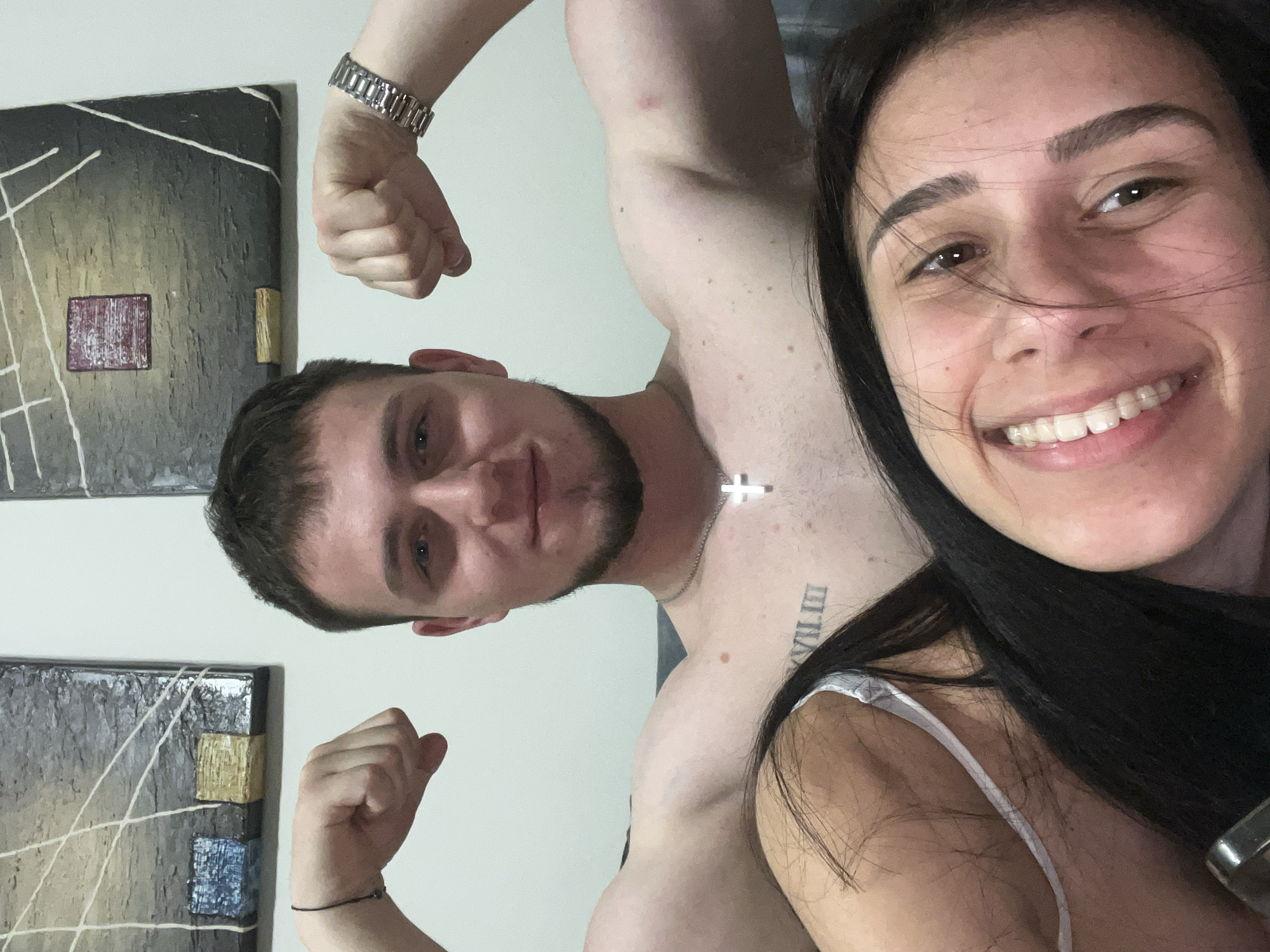 A selfie of a man and a woman. The man is on the left, shirtless and flexing his biceps. He has short brown hair and a beard, and is wearing a silver watch on his left wrist and a thin black bracelet on his right wrist. A silver cross necklace is around his neck and a tattoo in Roman numerals on his chest. The woman is on the right, with long black hair. She is smiling widely, and is wearing a white strappy top. Behind them are two pieces of abstract art hanging on a white wall. The scene takes place indoors. Unknown, Unknown.