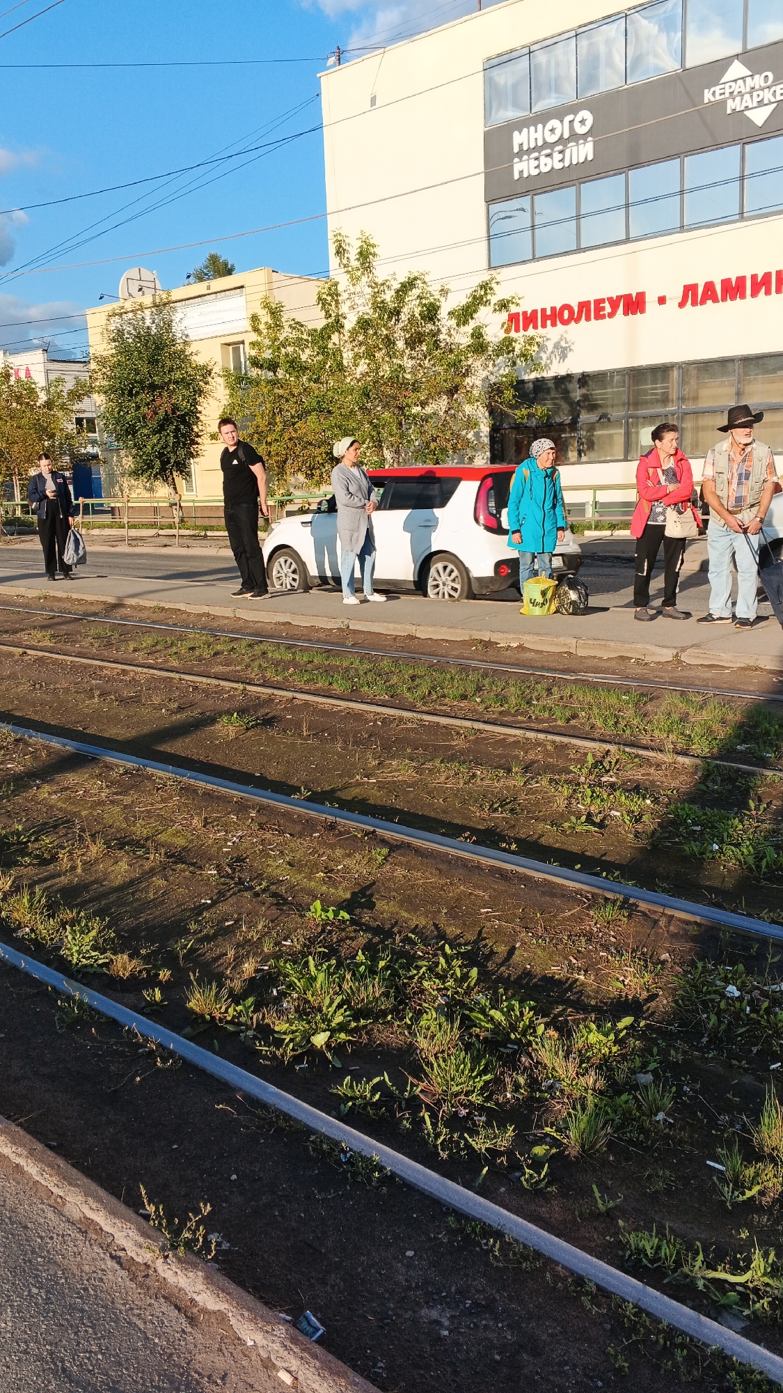 A group of five individuals is standing on a sidewalk next to a white car with a red stripe near a tram track. The car appears to be a Kia Soul. The building behind them has signage that reads "МНОГО МЕБЕЛИ" (Many Furniture) and "КЕРАМО МАРКЕ" (Ceramics Market). The location is confirmed to be Izhevsk, Russia, at the intersection of Lenin Street and Kalinin Street. The tram tracks are in the foreground, with dry grass and dirt. The sky is clear and blue, indicating a sunny day. The people appear to be waiting, possibly for public transport or a ride.