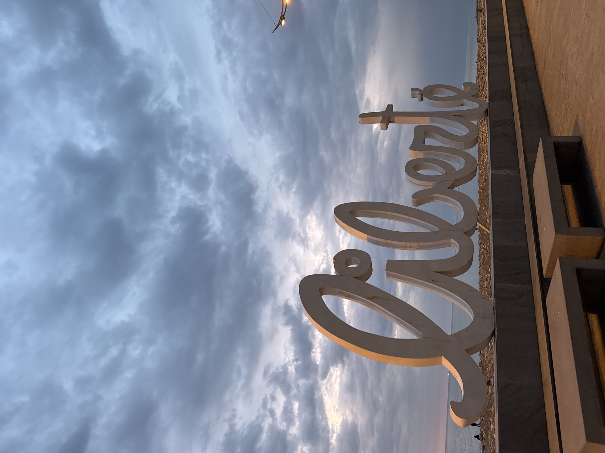The image captures a seaside scene under a cloudy sky. Dominating the foreground is a large, white, cursive-style sign spelling out what appears to be "Porto". The sign is mounted on a dark-colored structure that also features concrete blocks. The ground is covered with small pebbles or gravel. The ocean can be seen in the background, partially obscured by the sign. In the top left corner, there is a street lamp. The scene is at dusk or dawn, judging by the quality of the light. Two figures are seen in the background. The location is Porto, Portugal.