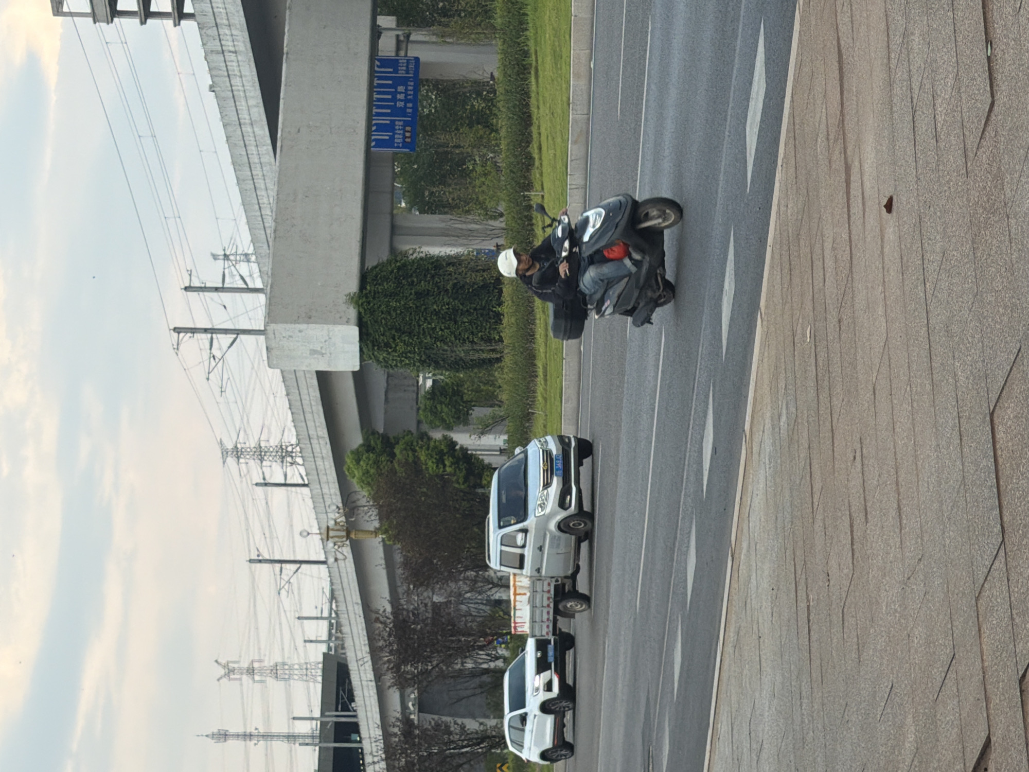 A scooter with a person riding it is traveling on a road in Jiangjin, China. The rider is wearing a white helmet and a dark jacket, and is riding on the right side of the road. Behind the scooter, a white SUV is towing a small trailer. The trailer appears to be carrying white rectangular objects. Further down the road, a light-colored van is also visible. Overhead, a concrete overpass supported by pillars is present, with power lines running across it. The sky is overcast with scattered clouds. The road is paved and has white lane markings. A sidewalk made of grey rectangular pavers is visible in the foreground.