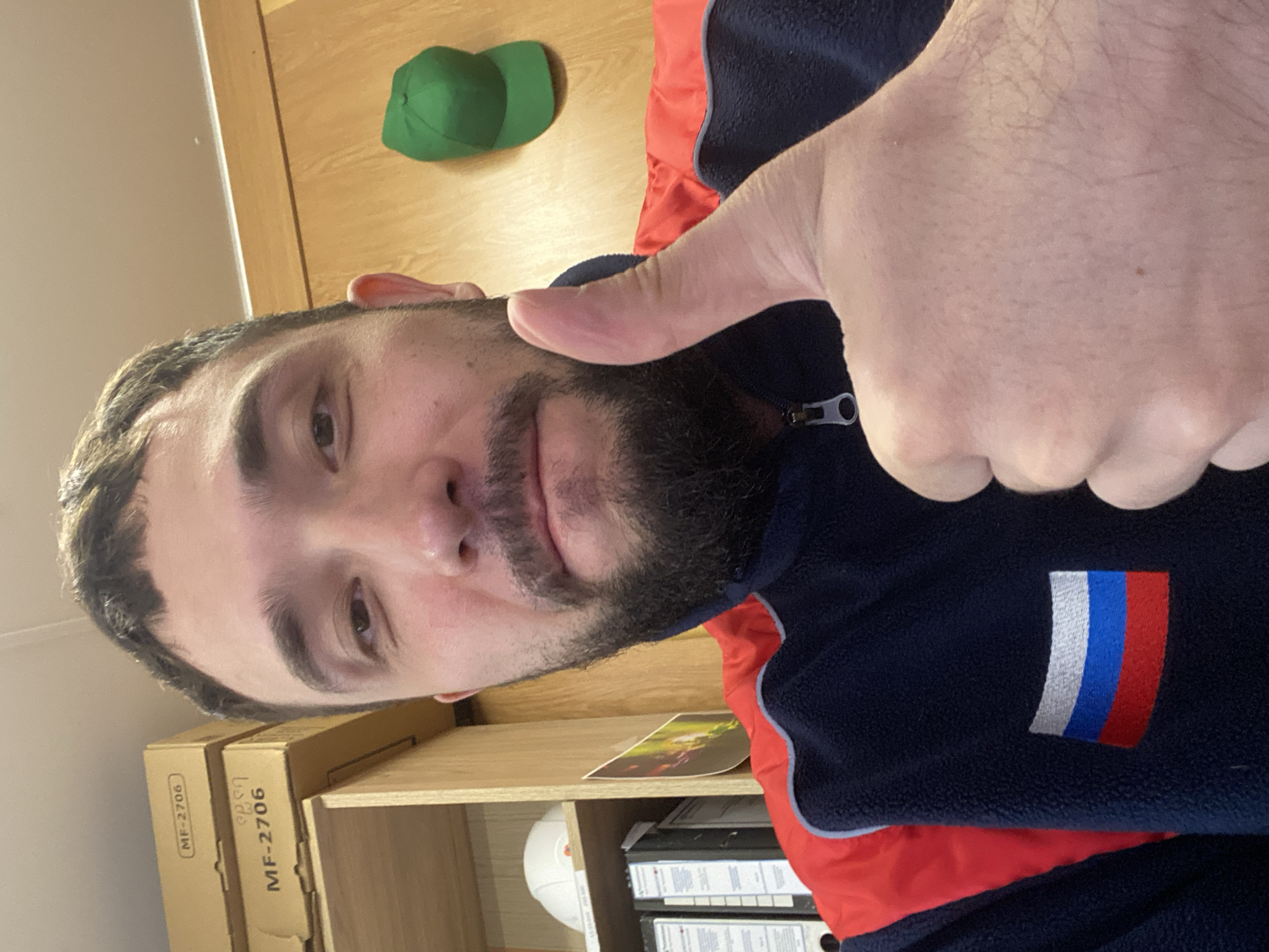A man with a beard is giving a thumbs-up gesture. He is wearing a navy blue and red jacket with a Russian flag patch. Behind him is a wooden shelf with boxes and a green hat. The location is not provided in the image.