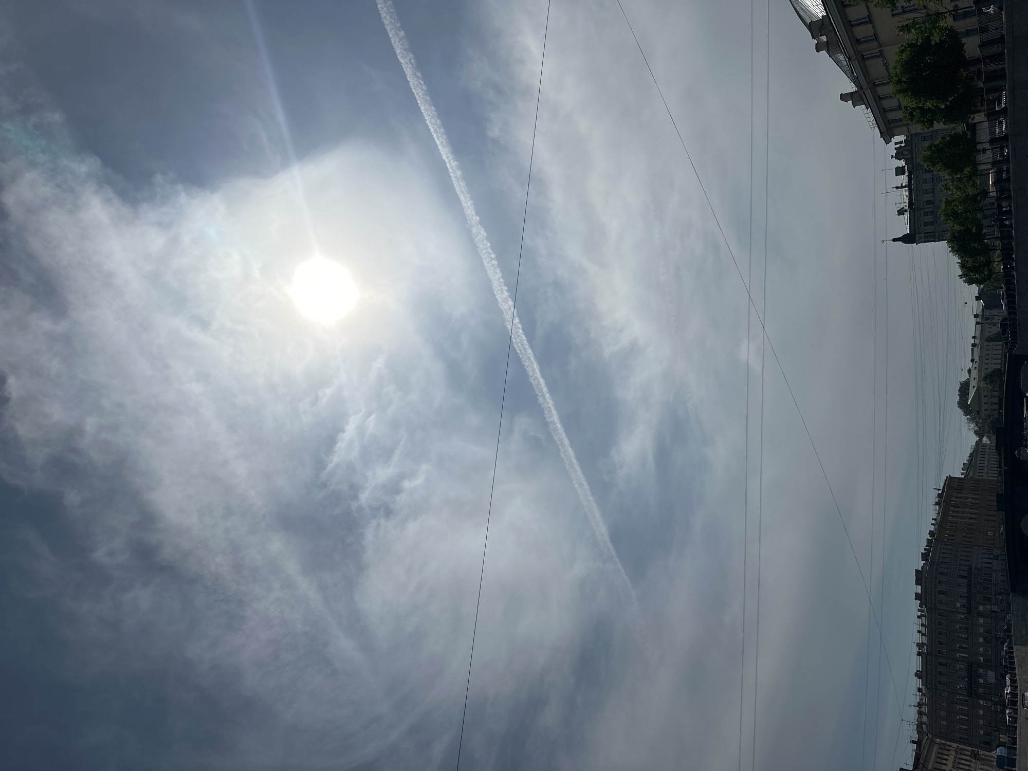 The image captures a street scene in Saint Petersburg, Russia, during daylight. The sky is predominantly visible, filled with wispy clouds and two prominent contrails left by airplanes. The sun shines brightly, obscured slightly by the cloud cover. In the lower right portion of the frame, buildings line the street, with trees interspersed along the sidewalk. Power lines stretch across the sky, adding to the urban landscape. The photograph is oriented vertically.