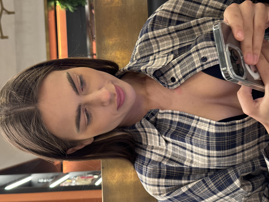 A female subject, appearing to be an adult, is depicted indoors in Verona, Italy, engaged with a smartphone. She has brown hair with a central part and is looking downward, focusing on the device held in both hands. She wears a blue and white plaid long-sleeved shirt, partially unbuttoned to reveal a black garment underneath. A small, erythematous mark is visible on her upper left chest. The smartphone is equipped with a clear protective case. The background features a golden-toned surface, vertical linear lighting elements on the left, and a horizontal orange-lit feature on the right, suggesting an indoor commercial or public space with artificial illumination. Green foliage is faintly visible on the far right.