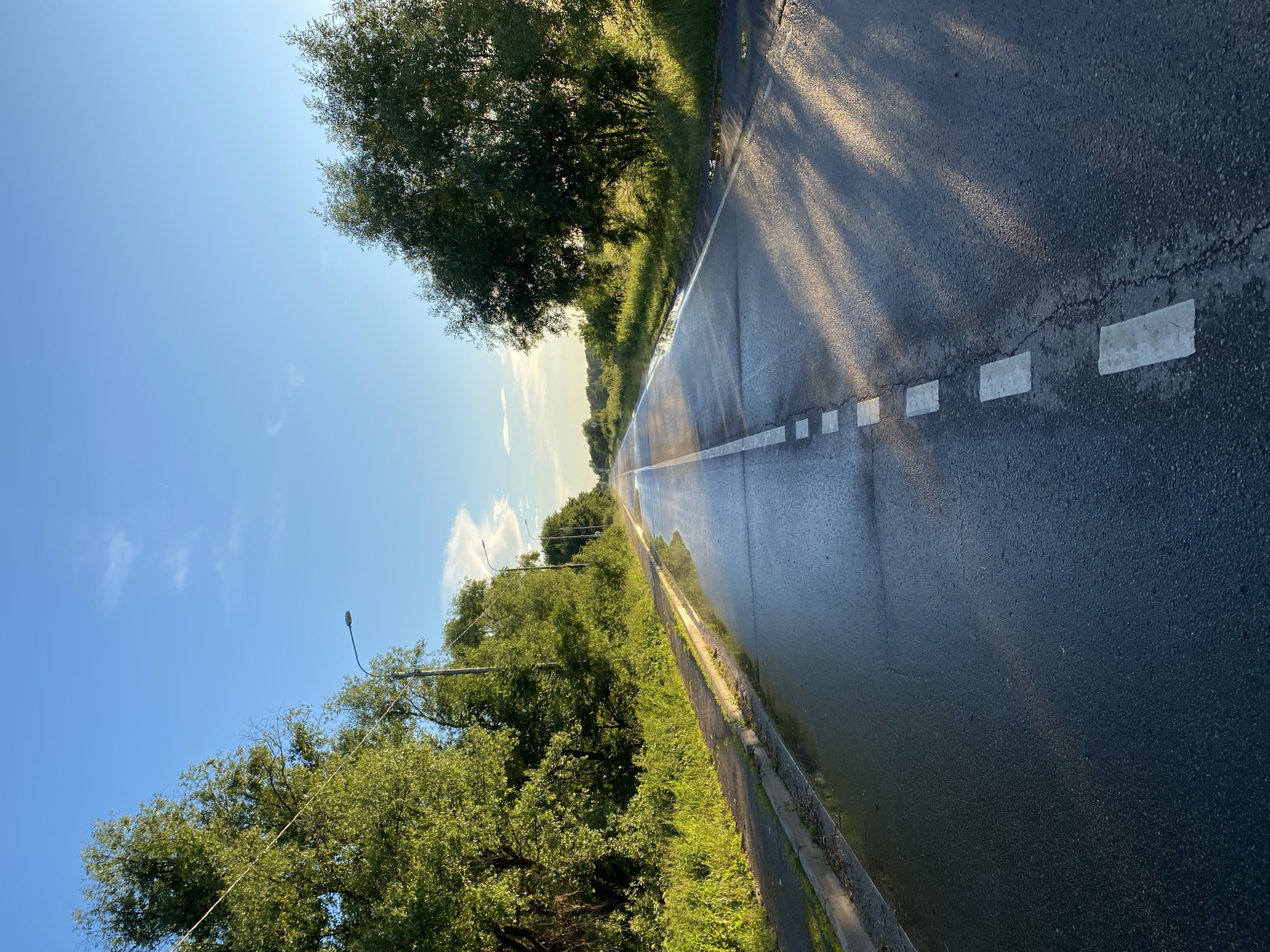 A wet, two-lane asphalt road stretches into the distance under a bright blue sky with scattered white clouds. The road is bordered by green trees and grass on both sides. Puddles of water reflect the sky and surrounding foliage on the left side of the road. White dashed lines mark the center of the road. Sunlight casts dappled shadows across the road's surface from the trees. Several streetlights are visible along the left side of the road. The scene appears to be in a rural or suburban area.