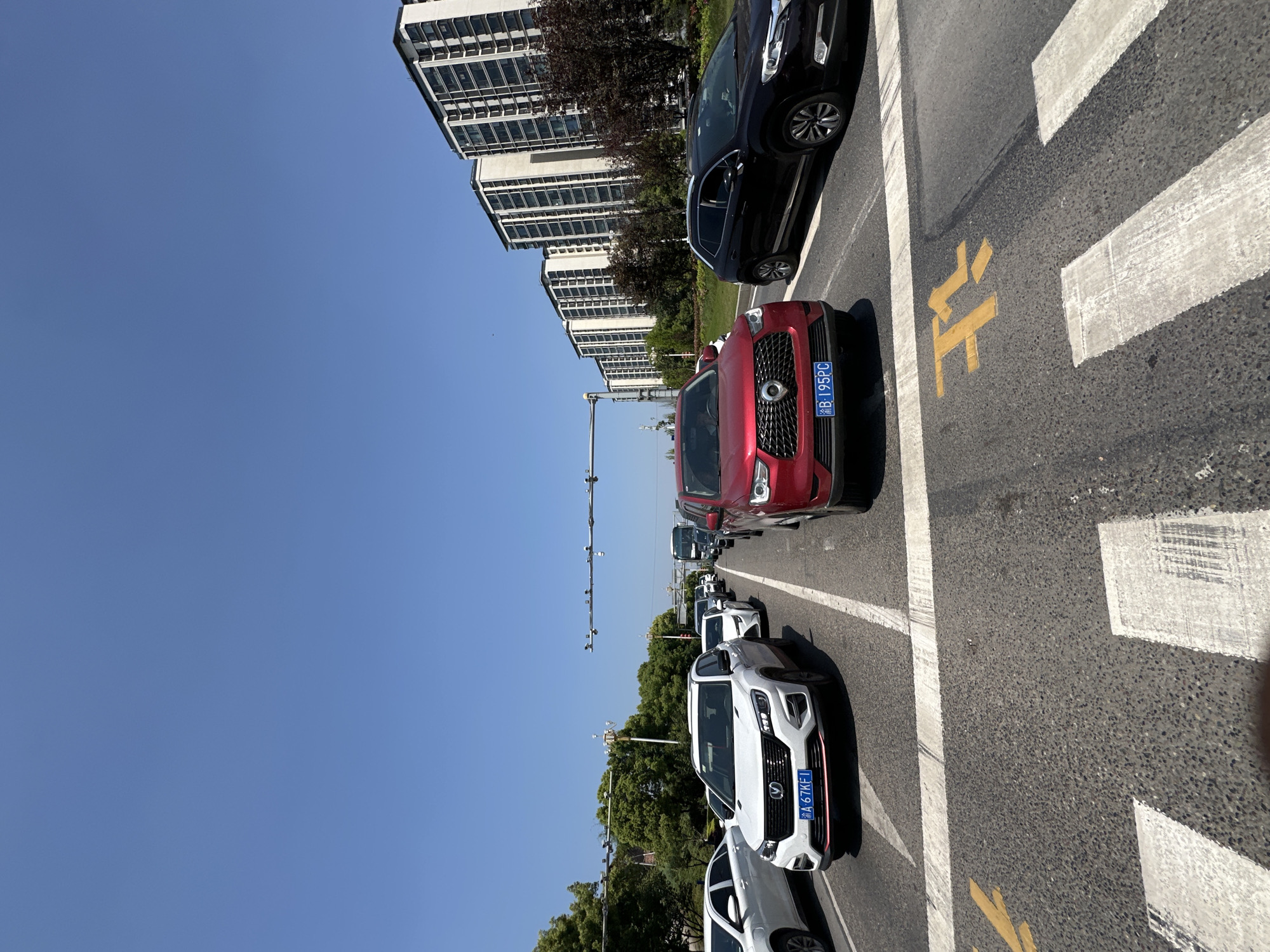 The image depicts a street scene in Jiangjin, China. Several cars are parked along the side of the road, including a red SUV with license plate "渝B·195PC" and a white SUV with license plate "渝A·67KF1". The road has pedestrian crosswalks marked with white stripes and yellow "wait" symbols. In the background, there are tall buildings and trees. The sky is clear and blue. There are traffic lights and surveillance equipment visible. No people are directly observable in the image.