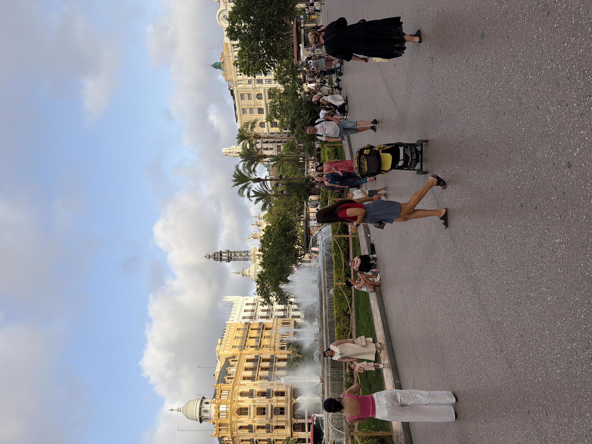 An outdoor, eye-level, full shot shows a street scene in front of an ornate building with a fountain and many people walking. The sky is cloudy, and the sun is shining through the clouds. The street is paved with asphalt. In the foreground, a woman in white pants and a pink tank top stands facing away from the camera, with her left side closer to the camera. To her left, a child in a pink dress walks to the left. A woman in a light beige dress stands to the left of the child. In the midground, a fountain with multiple tiers sprays water into the air. Several people are seated on the grass to the right of the fountain, and palm trees are visible behind them. The building in the background is yellow with white trim and features several arched windows and a dome on the left side. Another building to the right is white with green accents. People are walking and pushing strollers on the street. In the right midground, a woman in a black dress walks away from the camera.