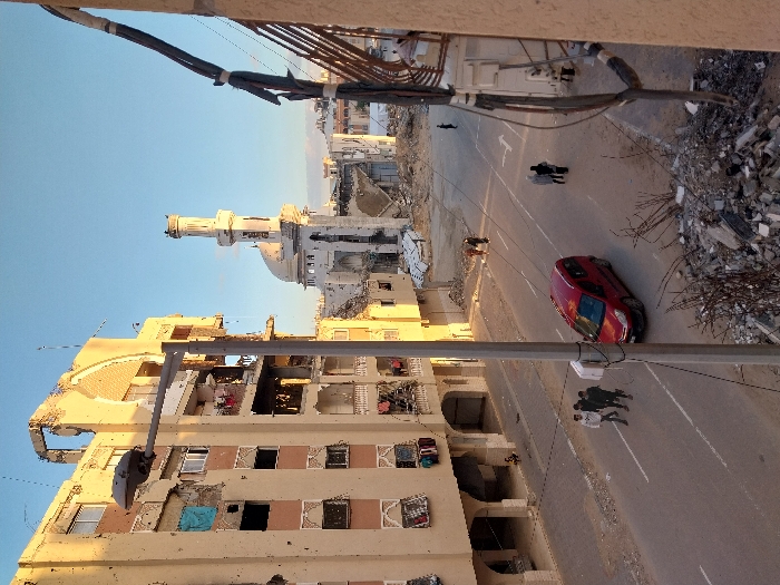 The scene depicts a street ravaged by conflict. Buildings exhibit severe damage, with missing sections and exposed interiors. Debris litters the roadside, indicating destruction. A red car is parked on the street. Several figures walk along the road. A mosque with a prominent minaret stands amidst the devastation, partially damaged. Overhead, power lines crisscross the sky. The scene suggests a war-torn urban landscape.