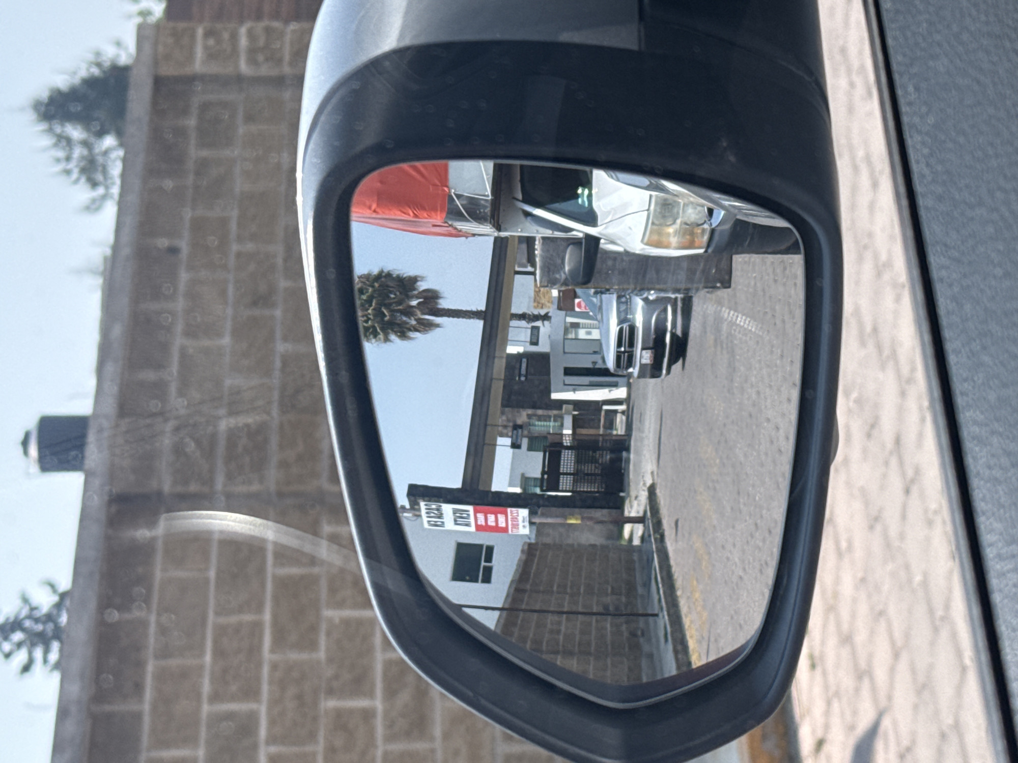 The image shows a side-view mirror reflecting a street scene in San Andrés Cholula, Mexico. In the reflection, a white pickup truck with a red cargo cover is closely followed by two other cars. A palm tree is visible on the left side of the reflected street. The building in the background has a sign in red and white that reads "GAS CIL EN." The street is paved with gray stone. Outside of the mirror, a brick wall is visible.