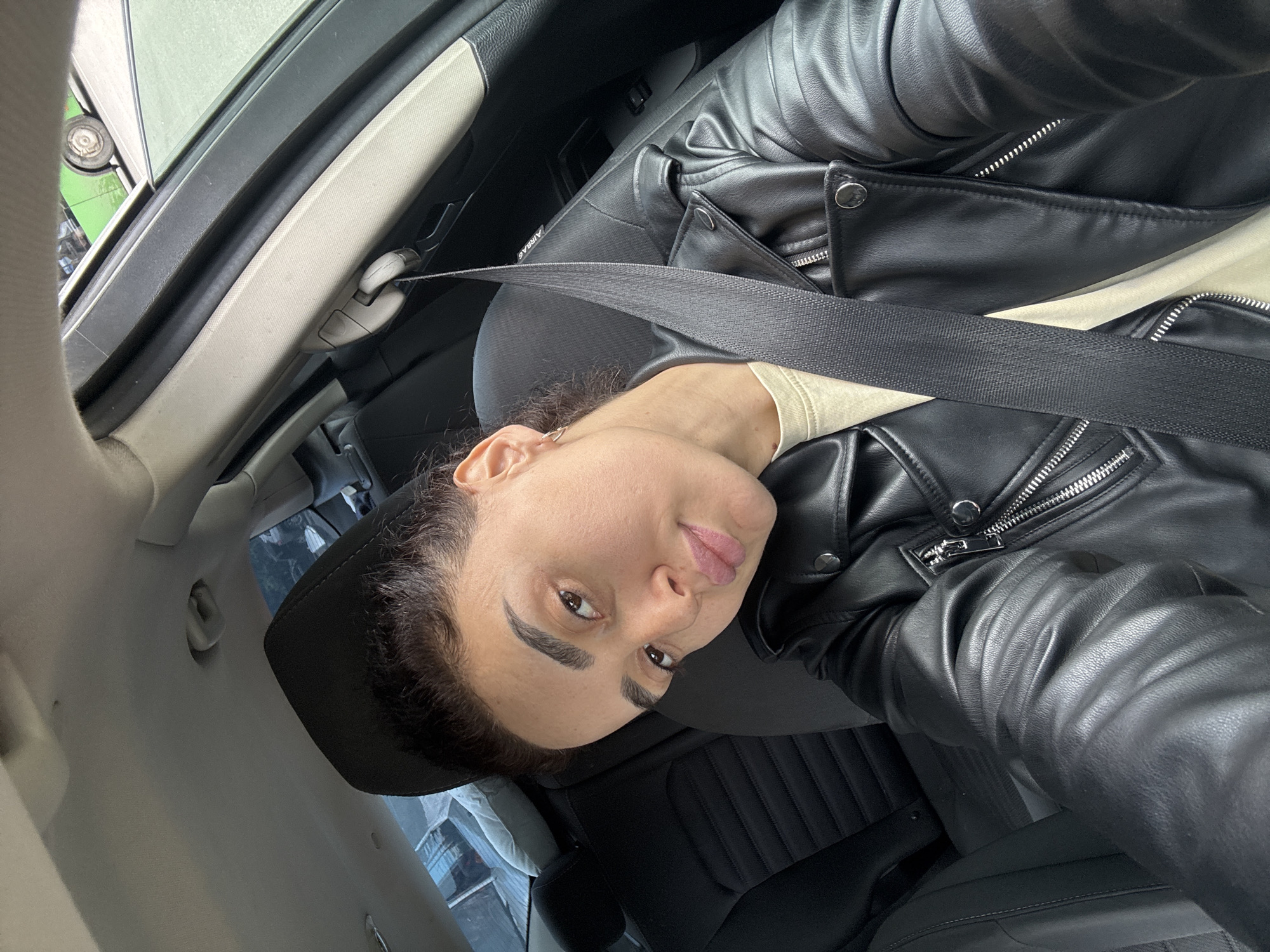 A woman in a black leather jacket is inside a car with her seatbelt on. The car appears to be parked or stopped. The interior is mostly dark-colored, with hints of light from outside. The location is inferred to be Tbilisi, Georgia, but there is no visual confirmation of the city.