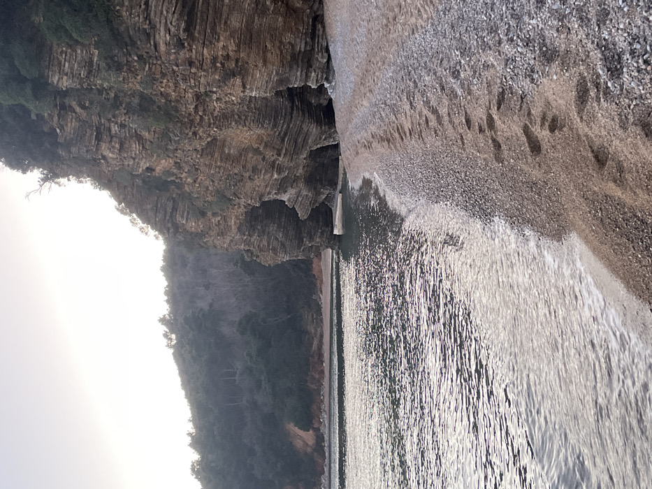 The scene depicts a pebble and sand beach along the Adriatic coast at Mogren Beach, Budva, Montenegro. The shoreline extends diagonally from the lower-left to the middle-right of the frame. The sea on the left is calm, exhibiting subtle ripples and reflecting a diffused light from a pale, hazy sky. Gentle waves create a thin line of white foam where they meet the shore. A series of distinct human footprints is present on the reddish-brown pebble and sand mixture of the beach, extending linearly into the background along the waterline. To the right, prominent stratified rock cliffs rise directly from the water's edge, characterized by distinct horizontal layering in earthy brown and orange tones. At least two large cave-like openings or deep overhangs are visible at the base of these cliffs. Sparse green vegetation clings to the cliff faces, and a dense forest of dark green trees covers the hillsides in the background, receding into the distance under the pale sky.