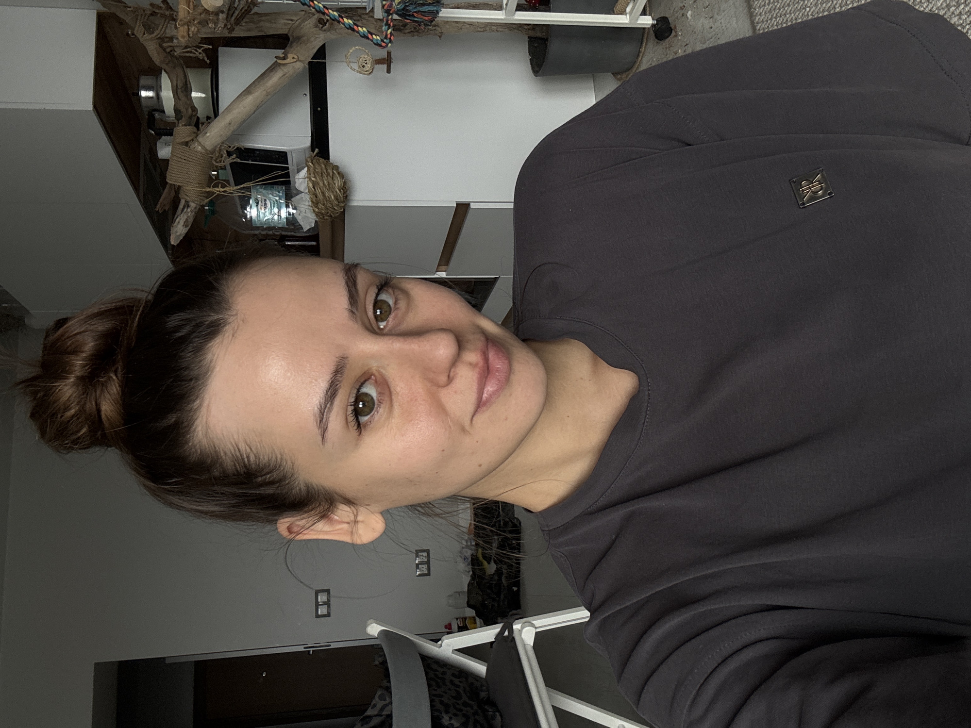 A portrait-style image, taken indoors in Krasnoyarsk, Russia, features a young woman. Her head and upper torso are visible, with her gaze directed towards the camera and a slight, pleasant smile on her lips. She has brown hair pulled back into a bun, fair skin, and warm brown eyes. She is wearing a plain dark grey long-sleeved sweatshirt. On the left side of her chest, a small, dark rectangular emblem with what appears to be stylized "JR" or similar initials is visible.
The background suggests a casual, domestic setting. To the woman's right, a complex wooden structure with various ropes and knots is prominent, possibly serving as a pet perch or a decorative piece. A clear plastic bottle with the faint text "НАРЗАН" is visible nestled within this structure. Further in the background, a dark grey plant pot, shelves or cabinets with white and grey surfaces, and what looks like a white clothes drying rack are discernible. The lighting is moderate, typical of an indoor environment.