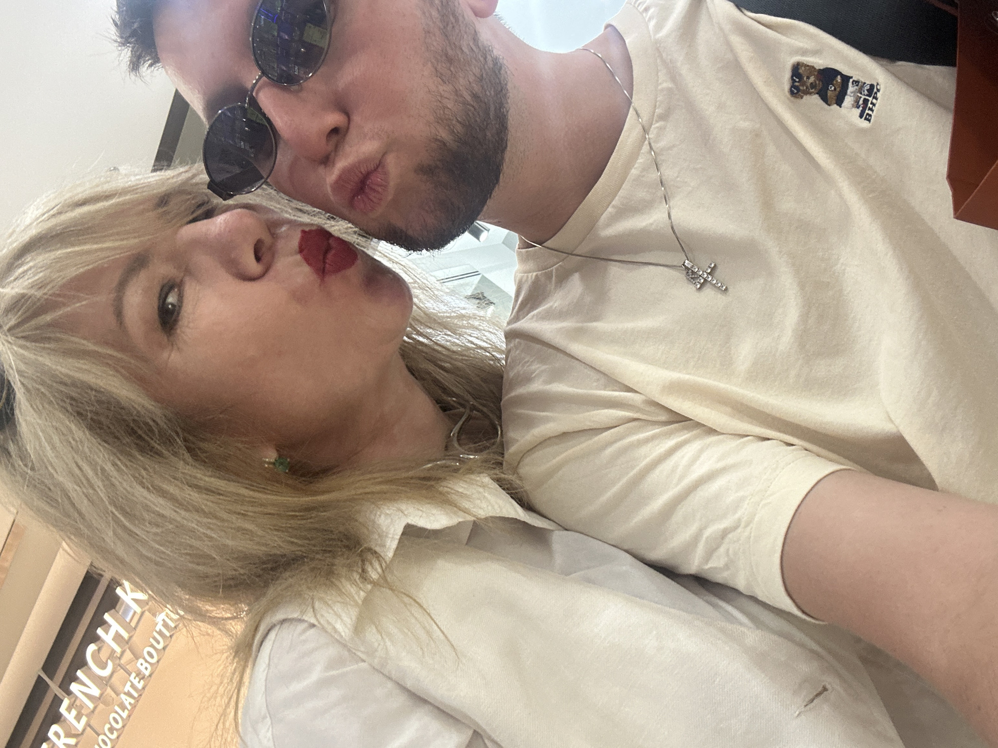 A close-up selfie shows a fair-skinned woman with blonde hair and red lipstick puckering her lips toward the camera. She wears a white shirt and a green earring. To her right, a fair-skinned man with short brown hair and a light beard puckers his lips. He wears a light-colored t-shirt with a silver cross necklace and a small embroidered bear logo. He also wears dark, oval sunglasses. The background appears to be indoors, with a sign that reads "FRENCH K CHOCOLATE BOUTIQUE." The lighting is bright and suggests an indoor commercial setting. The image does not provide enough visual information to determine the city of Moscow, Russia.