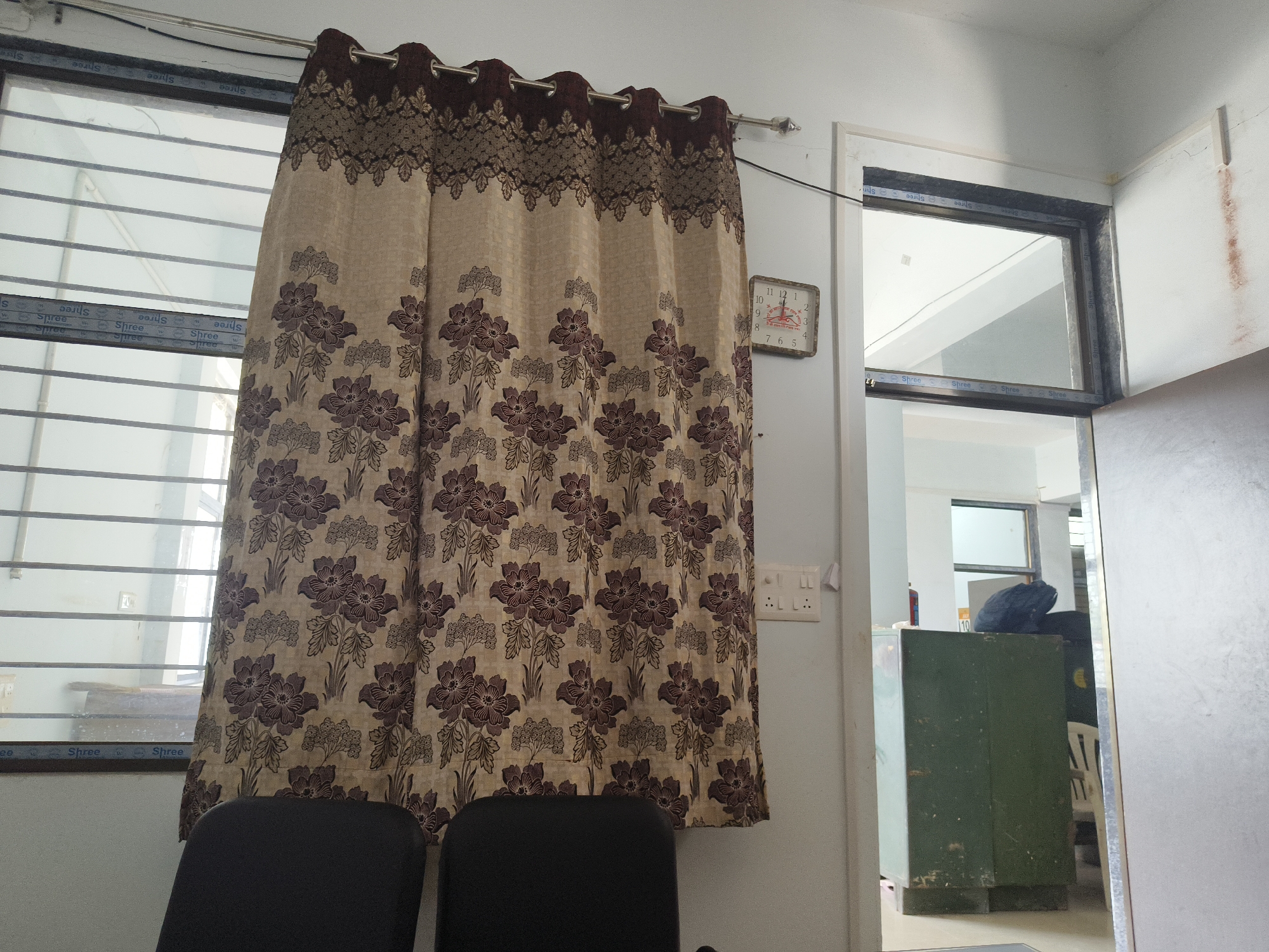 The image shows an indoor scene, likely a room in Dhandhuka, India. In the foreground, two dark-colored chairs are partially visible. A patterned curtain, with a beige background and floral motifs in brown and darker beige, hangs over a window that has horizontal bars. To the right of the curtain, a square clock with Roman numerals is mounted on a light-colored wall. Further to the right, a doorway leads to another area where a dark green metal cabinet is visible. A dark backpack rests on top of the cabinet. The overall environment appears to be an office or a similar functional space.