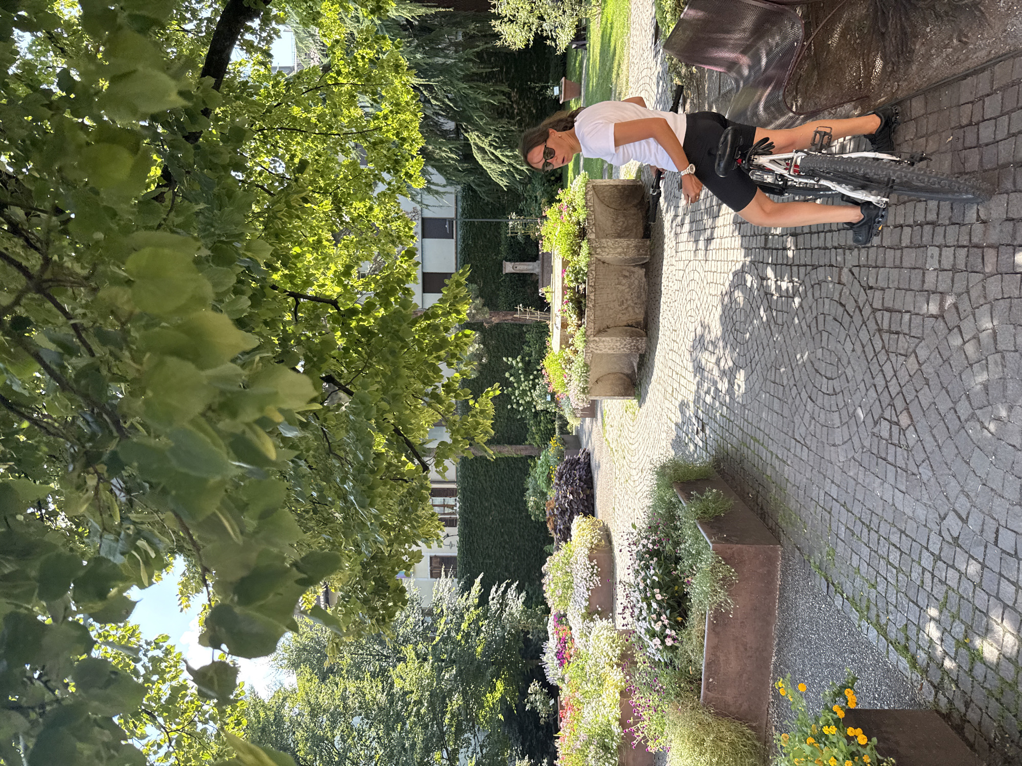 A woman wearing a white t-shirt and black cycling shorts is sitting on a bicycle in a public garden in the city of Trento, Italy. The garden features cobblestone pathways, raised planters filled with colorful flowers, and lush green trees. In the background, a building with ivy-covered walls and windows is visible. The woman is looking to her left, with a bench to her right. The sun is shining, casting shadows across the scene.
