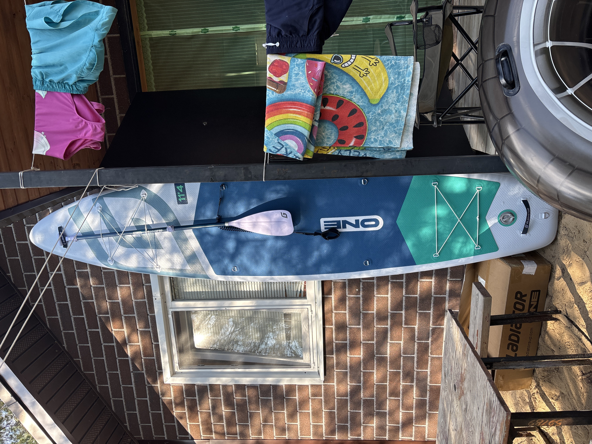 In the city of Kzyl-Bairak, Russia, a blue and white paddleboard stands upright against a brick building. A paddle is attached to the paddleboard. Nearby, on a clothesline, hang pink and blue swim bottoms and a colorful towel with fruit designs. To the left, a wooden table sits on sand. To the right, a foldable chair is placed next to the paddleboard. Partially visible in the foreground is a dark inflatable ring. The scene is outdoors, with sunlight casting shadows.