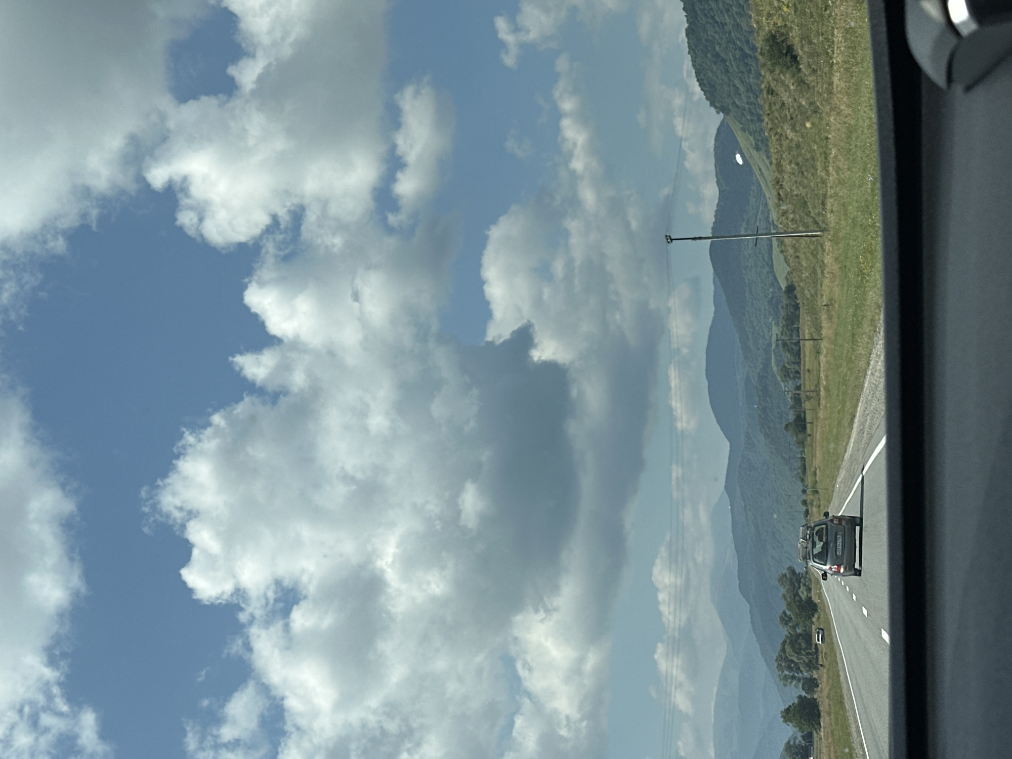 The image depicts a scene from a car driving on a highway. The road stretches ahead, flanked by grassy fields and trees, with a range of rolling green mountains in the background. The sky is a vibrant blue, filled with large, fluffy white clouds. A grey car with a roof rack is traveling in the same direction on the highway, in front of the camera vehicle. The interior of the car from which the photo is taken is visible at the bottom of the frame, showing part of the dashboard and windshield. Power lines and poles are also present along the side of the road. The location is identified as Nizhnyaya Yermolovka, Russia.