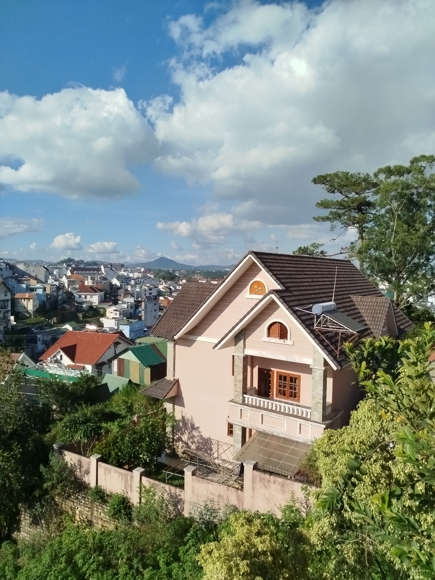 The image shows a three-story house with a brown tile roof in Da Lat, Vietnam, surrounded by greenery and other buildings. The house is light pink with a stone facade on the second story. It has a balcony with wood-framed windows and a small glass awning at the ground level. A solar water heater is installed on the roof. The background reveals an urban landscape with a mixture of buildings and a distant mountain. The sky is blue with scattered clouds. A concrete wall borders the property. The environment is lush, with trees and foliage in the foreground and around the house.