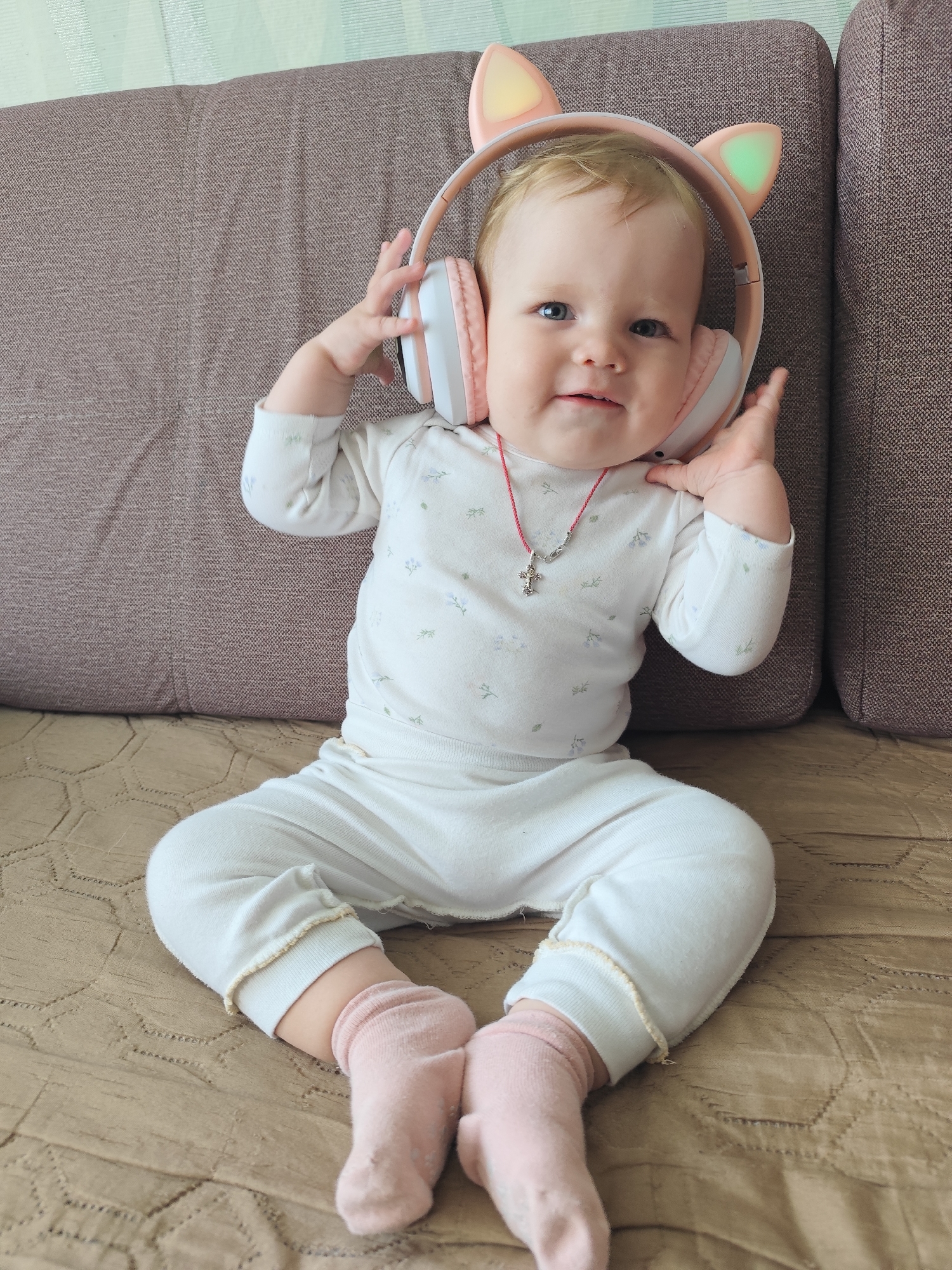 A toddler with fair skin, light hair, and blue eyes sits on a brown textured surface, likely a bed or couch. They are wearing a white long-sleeved onesie with a subtle floral pattern, pink socks, and a red cord necklace with a silver cross pendant. The toddler has headphones with illuminated cat ears on their head and is holding the headphones with their hands. The background consists of a brown couch. The image is a close-up, full-body shot of the toddler. The location in the city of Фоки, Russia cannot be determined from the image.