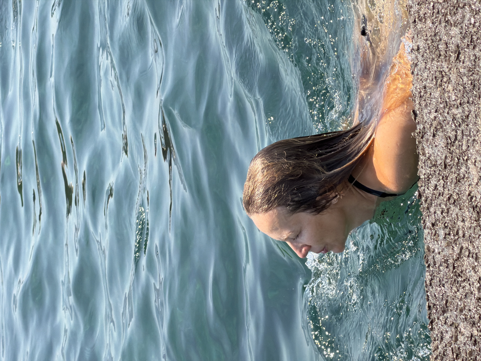 In Budva, Montenegro, a person with blonde hair, wearing a black bikini top, swims in the clear, blue water of the Adriatic Sea. The person is positioned in the middle of the frame, partially submerged, with water splashing around their head and shoulders. The foreground of the image features a textured, rocky surface, likely a beach or pier. The background is dominated by the rippling surface of the sea, with sunlight glinting off the water.