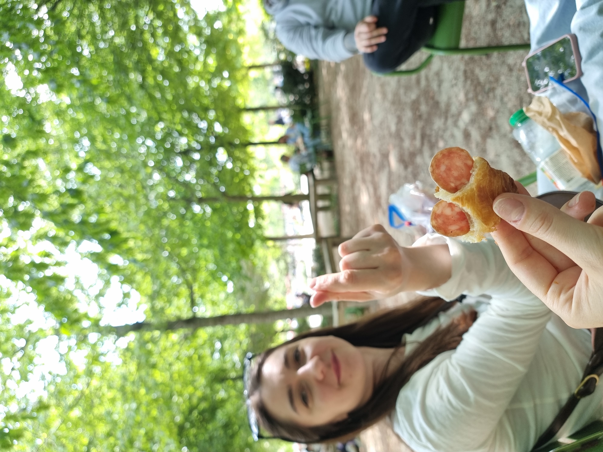 Here is a detailed description of the image:
The image is taken outdoors, likely in a park setting, given the background of lush green trees, which appear as a bokeh effect in the upper left. A young woman is in the foreground, holding a sausage roll. She has long brown hair, is wearing sunglasses perched on her head, and is dressed in a white shirt. The woman is looking at the camera with a slight smile.
In the middle ground, there are other people. One person is partially visible, seated on a green chair with their legs crossed. Further in the background, people are sitting on benches.
The foreground also includes a picnic setup. A plastic water bottle, a paper bag, and a mobile phone are visible on a table or the ground next to the woman.
The scene seems to be a casual picnic or gathering of friends in a park. Considering the park setting, the location could be one of the well-known parks in Paris, France, such as Jardin du Luxembourg, Parc des Buttes-Chaumont, or Jardin des Tuileries, but without specific contextual clues, it's difficult to pinpoint the exact location.