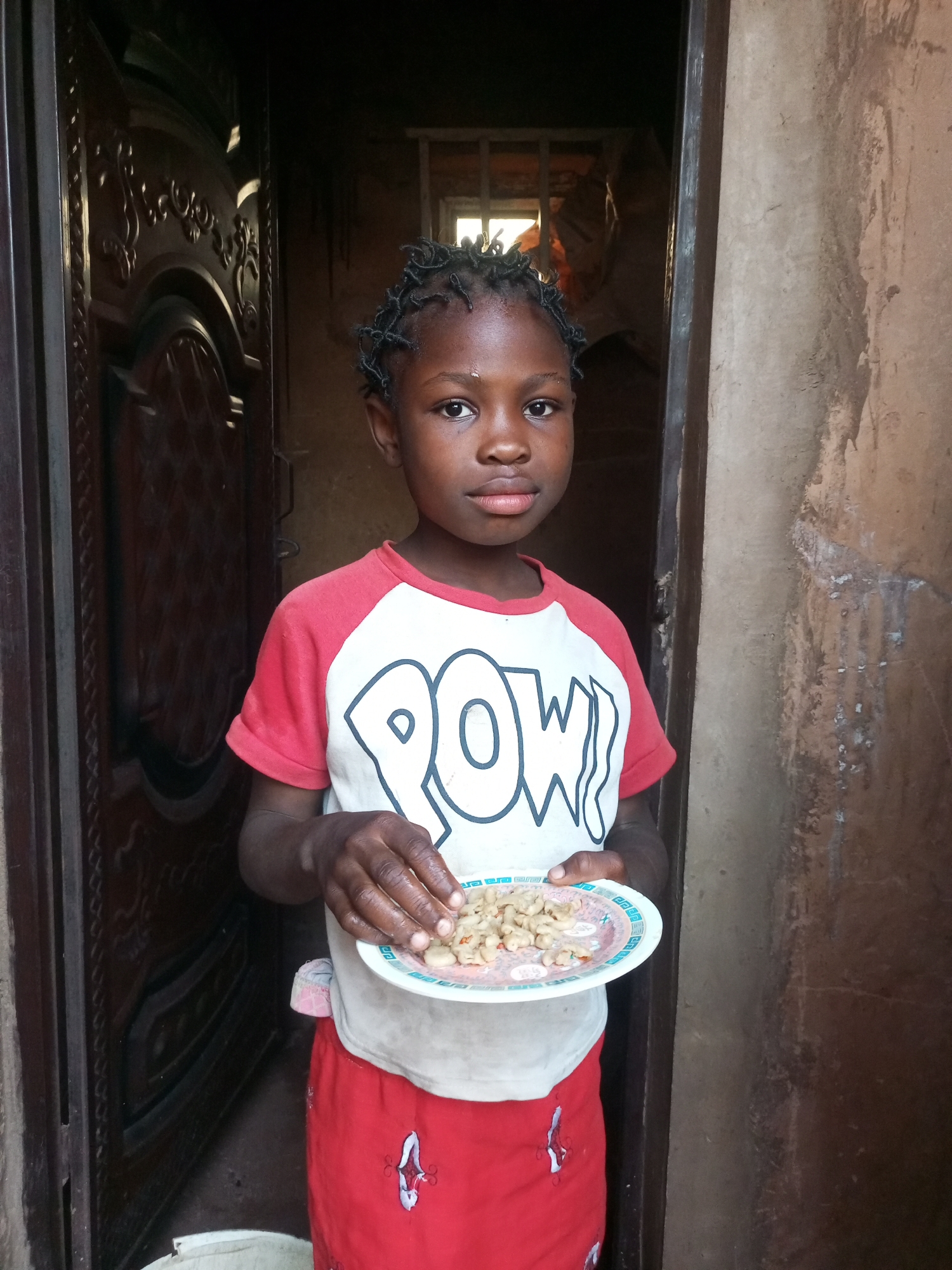 In an outdoor, dimly lit environment, a young Black child stands in a doorway, holding a plate of food. The child, with braided hair and wearing a red and white shirt with the word "POW!" printed on it and a red skirt, looks directly at the camera. The background reveals a dark, textured wall to the right and a large, ornate dark wooden door to the left. Sunlight streams in from a window in the background, illuminating the food on the plate. The food appears to be small, pale pieces, possibly pasta or legumes. The overall scene suggests a candid moment, perhaps in a residential area, with the child appearing to be posed for the photograph.